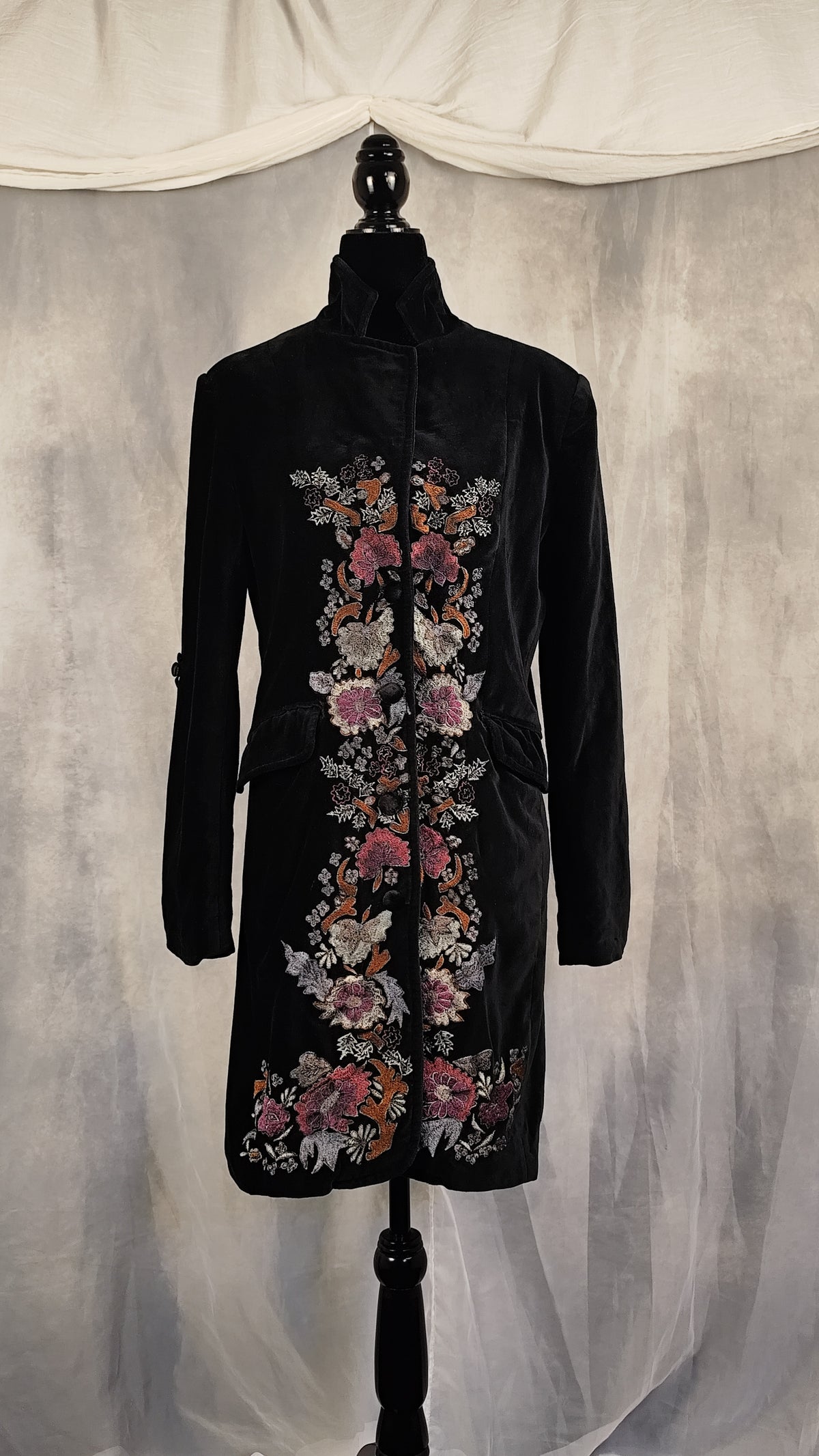 A black velvet longline coat with intricate floral embroidery in pink, orange, and white, displayed on a black mannequin against a draped gray fabric backdrop.