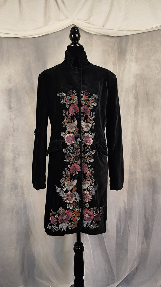 A black velvet longline coat with intricate floral embroidery in pink, orange, and white, displayed on a black mannequin against a draped gray fabric backdrop.