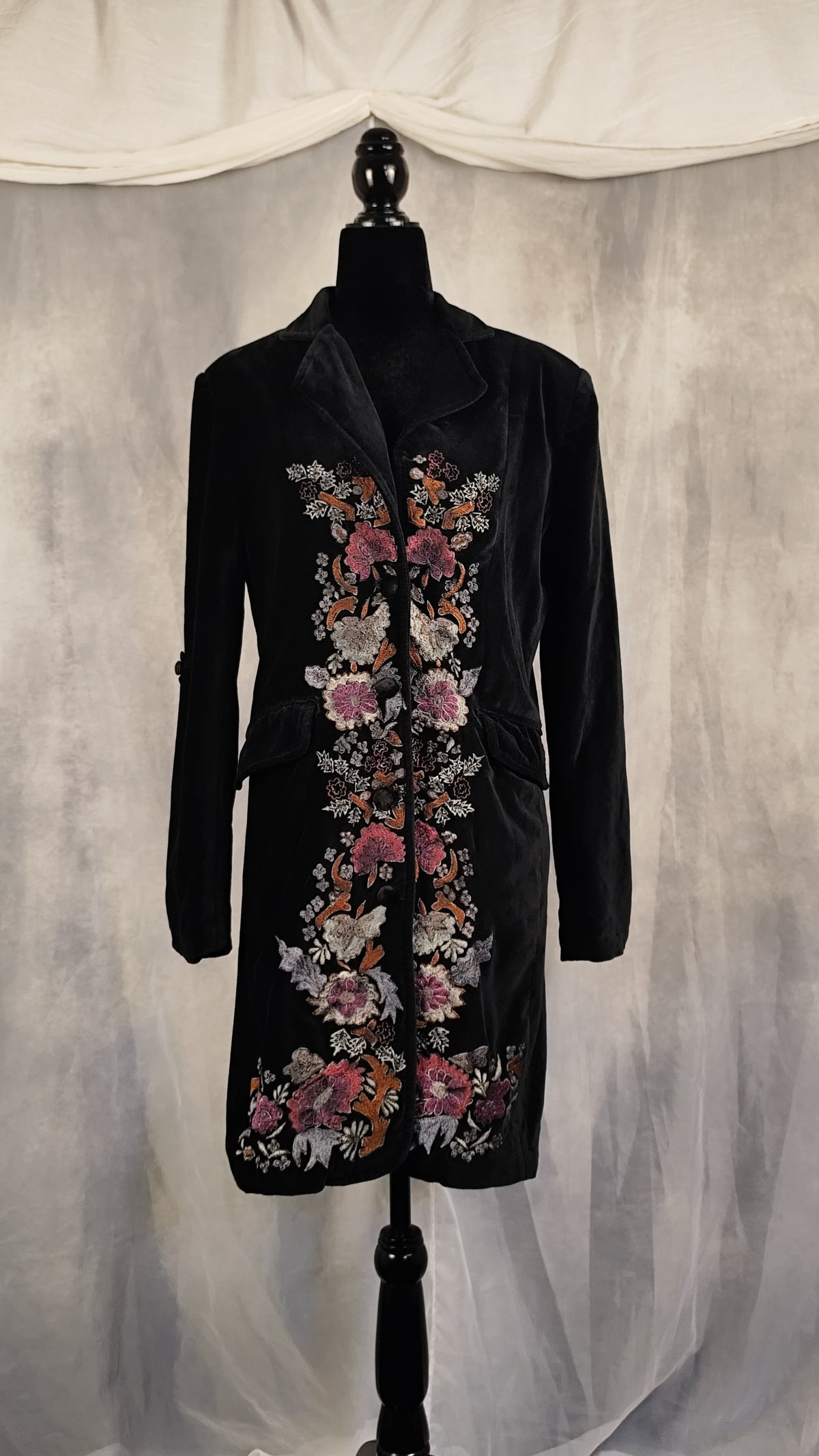 A black velvet longline coat with colorful floral embroidery, displayed on a black mannequin against a draped gray fabric backdrop.