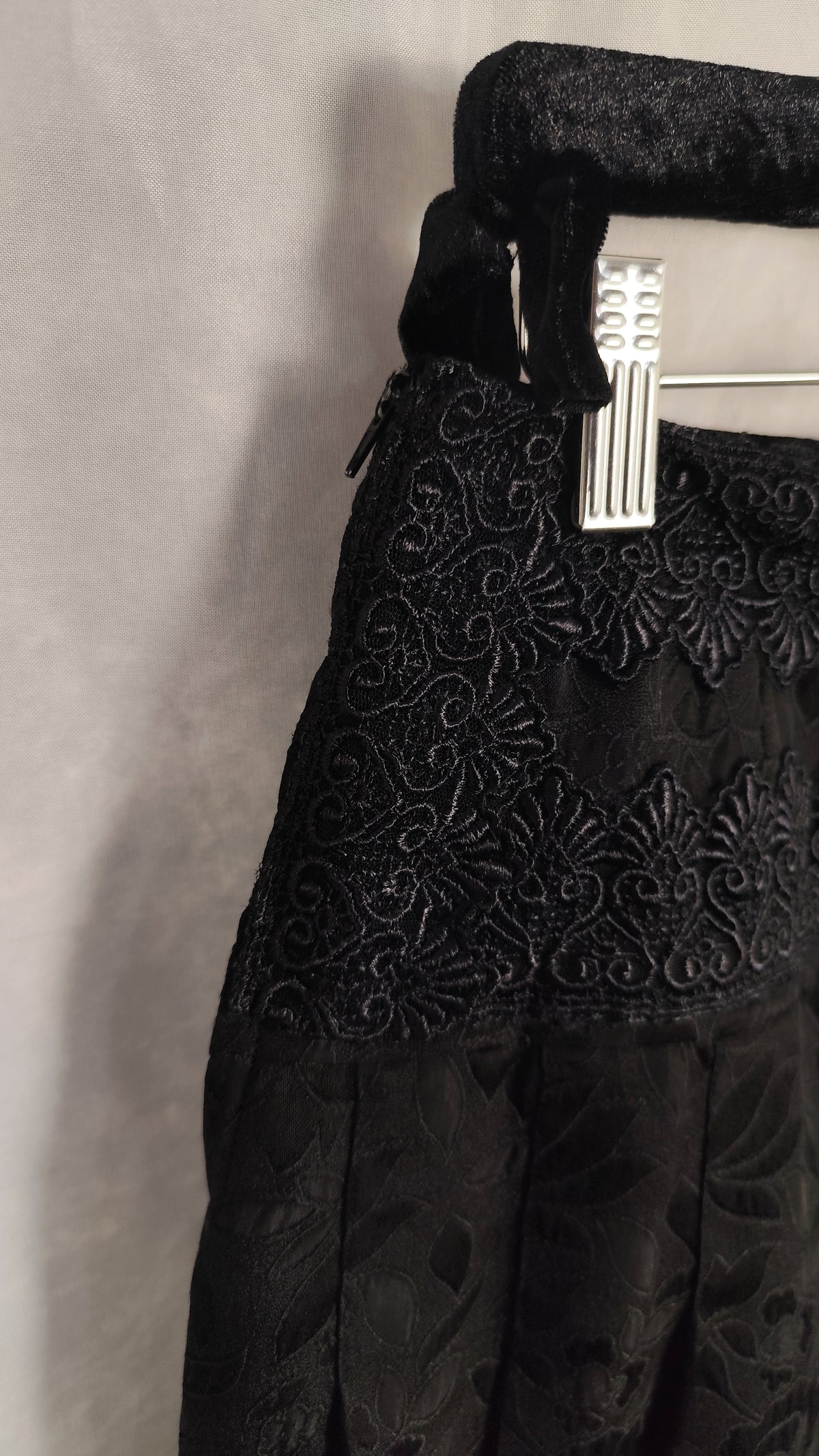A close-up of a black embroidered lace skirt or dress section hanging on a white plastic hanger against a light gray fabric backdrop.