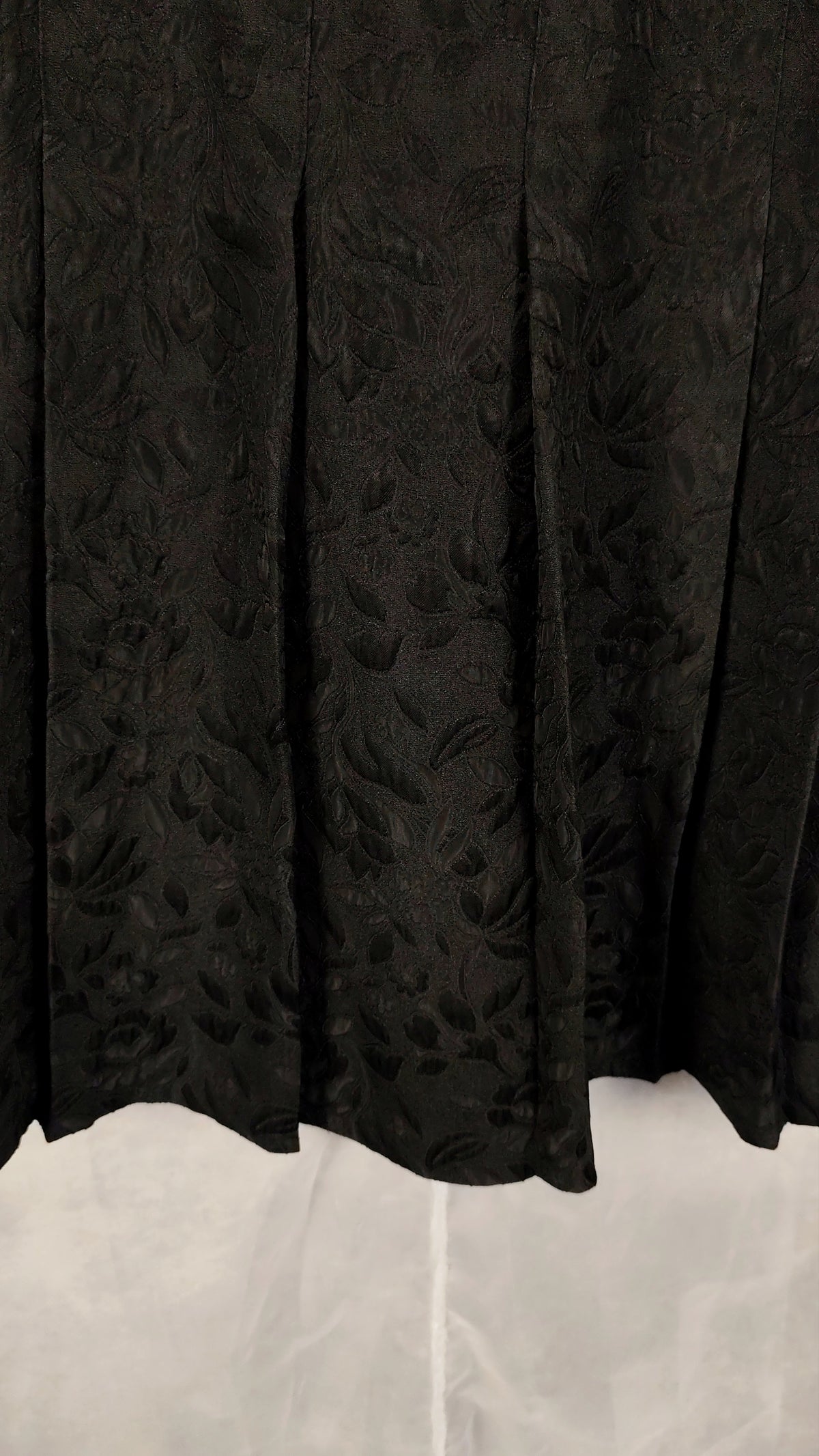 Close-up of a black pleated skirt with a subtle floral jacquard pattern, hanging against a light gray fabric backdrop.