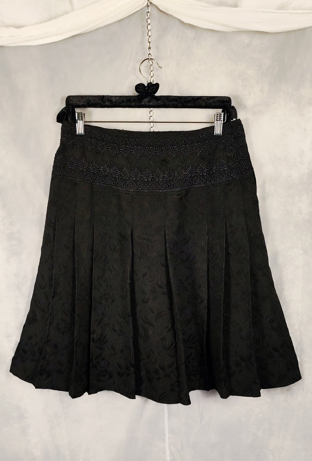 A black pleated skirt with a textured jacquard pattern and a decorative embroidered waistband, hanging on a black hanger against a light gray fabric backdrop.