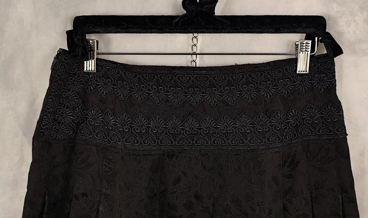 A close-up of a black skirt with intricate floral lace embroidery across the waistband, hanging from a black velvet hanger with silver metal clips against a textured gray background.