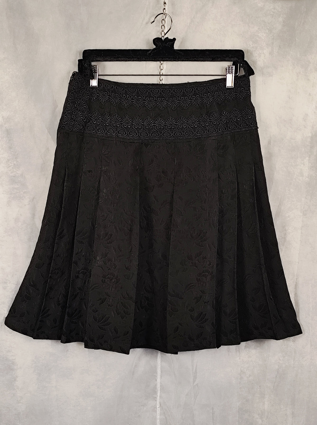 A black pleated skirt with a textured floral jacquard pattern and a lace-like waistband, hanging on a black hanger against a gray textured wall.