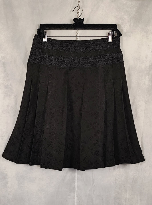 A black pleated skirt with a textured floral jacquard pattern and a lace-like waistband, hanging on a black hanger against a gray textured wall.