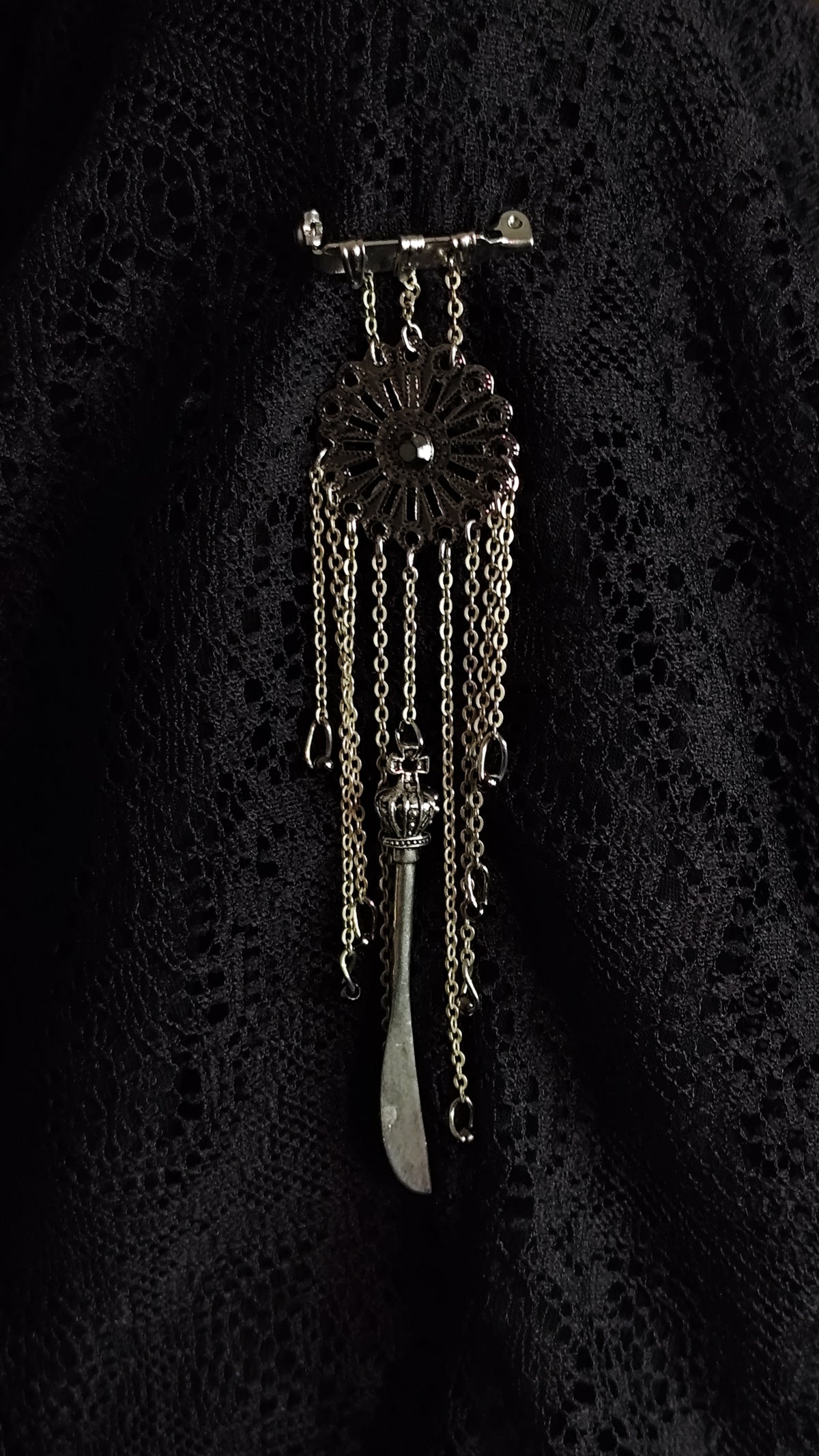 A vintage-style silver-toned brooch featuring a circular sunburst design with dangling chains and a teardrop-shaped charm, displayed on a textured black lace fabric background.
