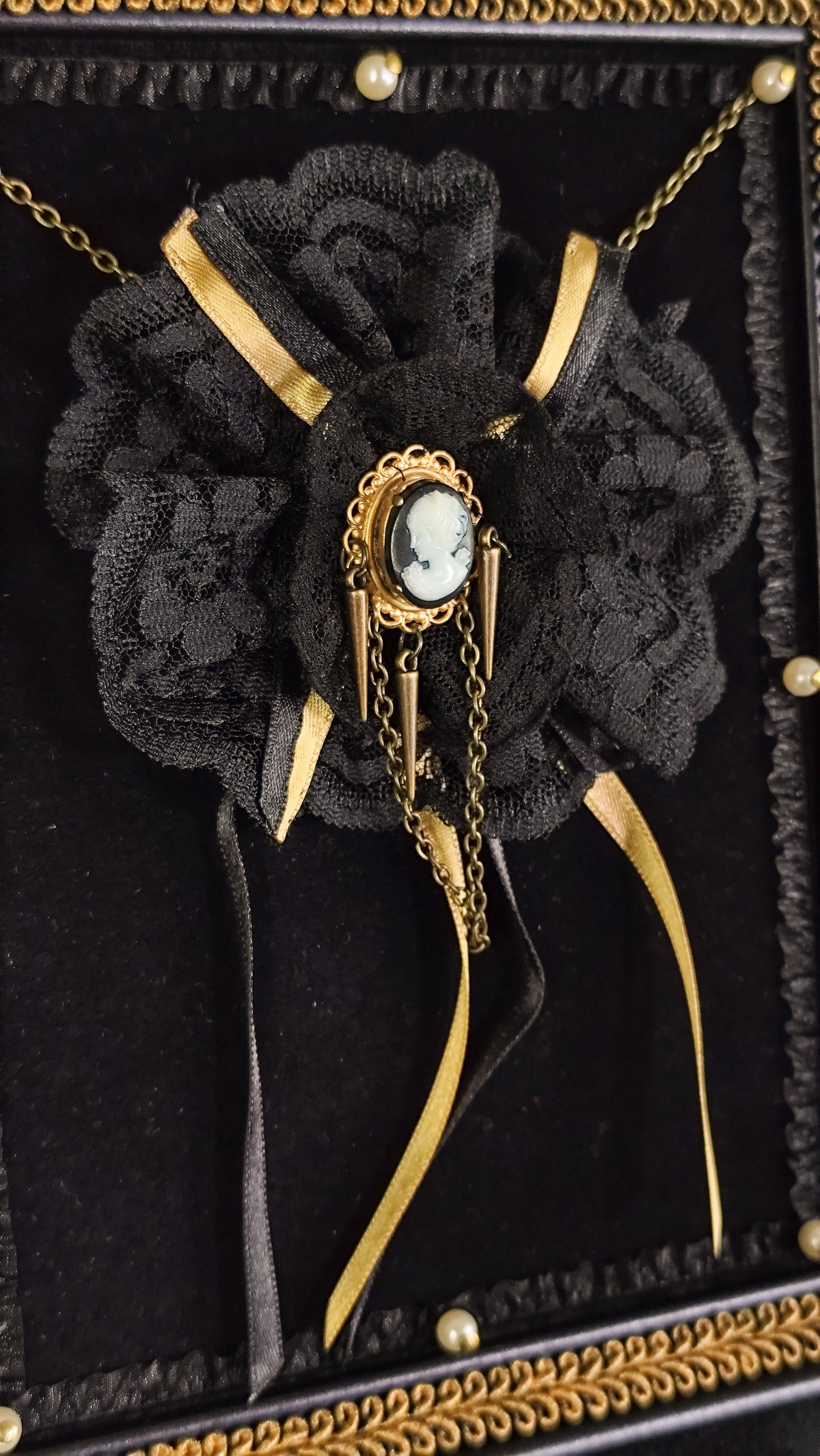 A black lace rose brooch with gold ribbon accents and a central oval plastic cameo, displayed on a black velvet jewelry box with gold trim.
