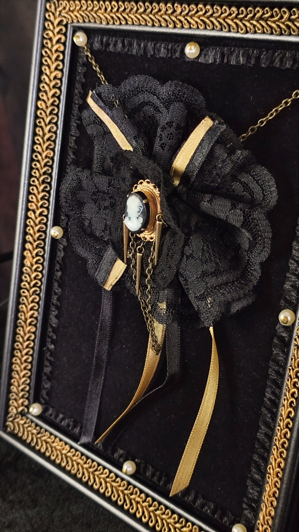 A gothic-style black lace flower brooch with gold ribbon accents and a central cameo, displayed inside a black velvet shadow box with an ornate gold frame.