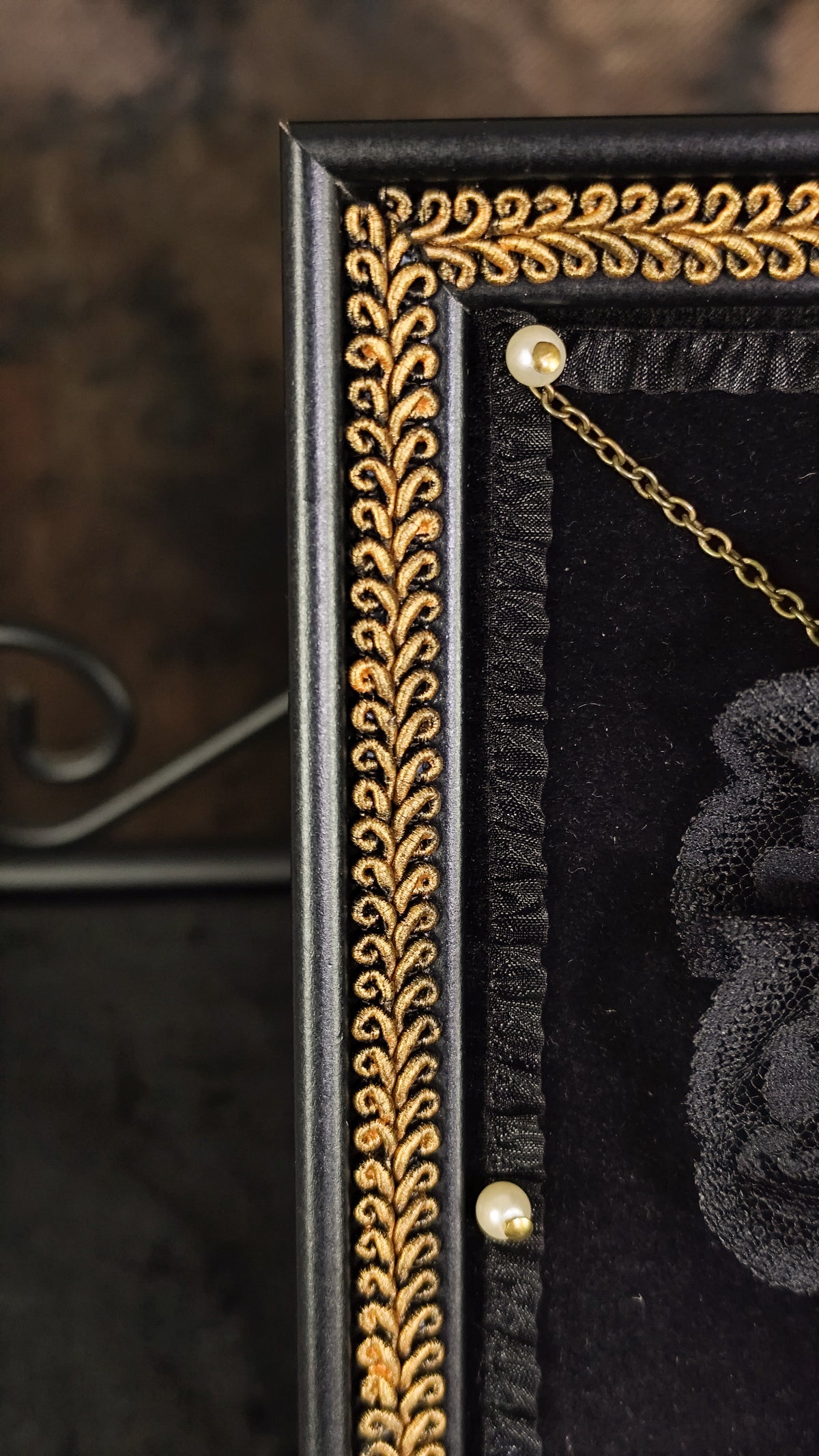 Close-up of a black velvet display case with an ornate gold decorative border, a gold hanging chain, and a black embroidered rose detail.