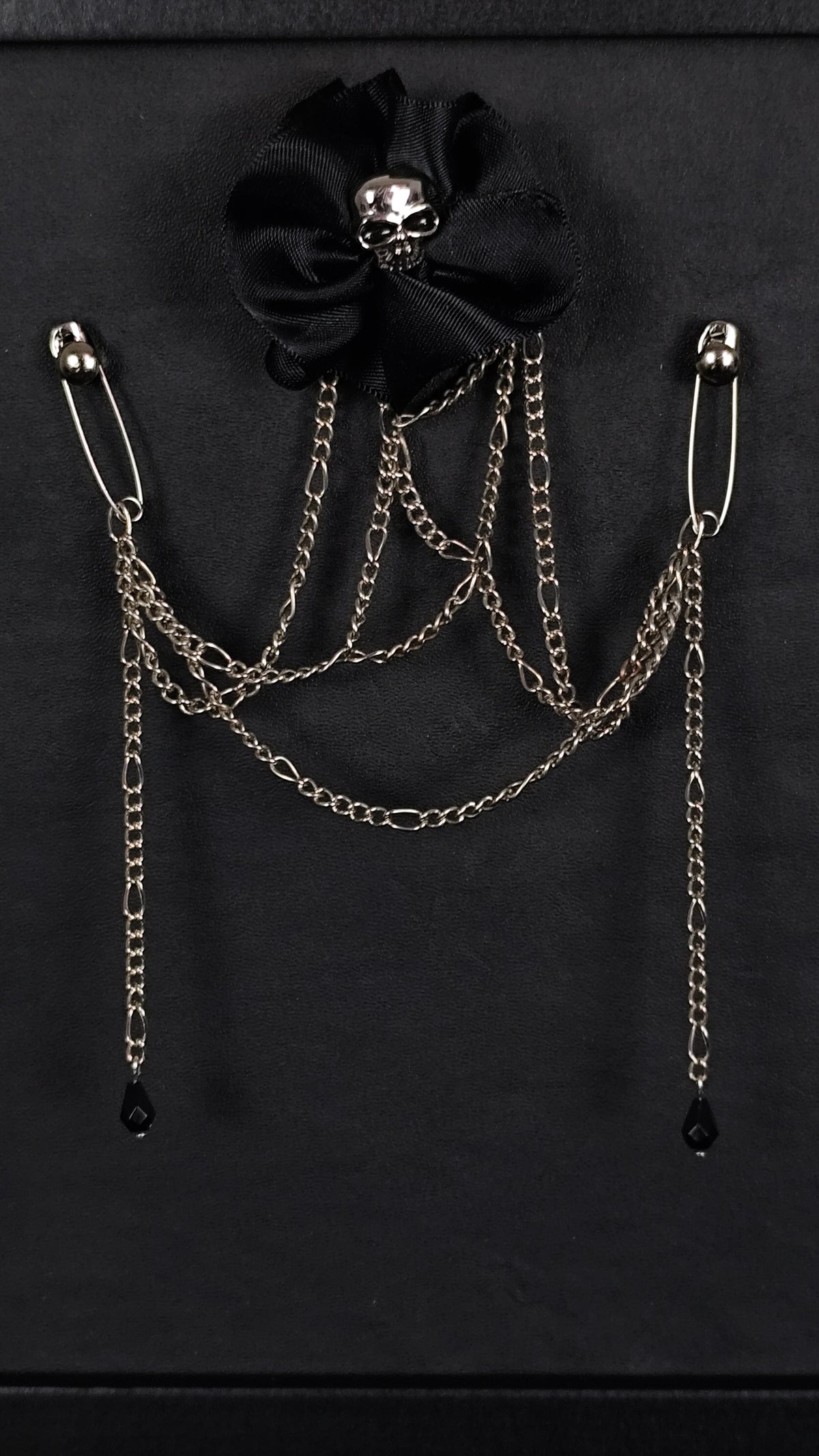 Black gothic brooch featuring a black fabric flower with a silver skull center, adorned with draped silver chains and safety pin ends terminating in small black teardrop charms, displayed on a black background.