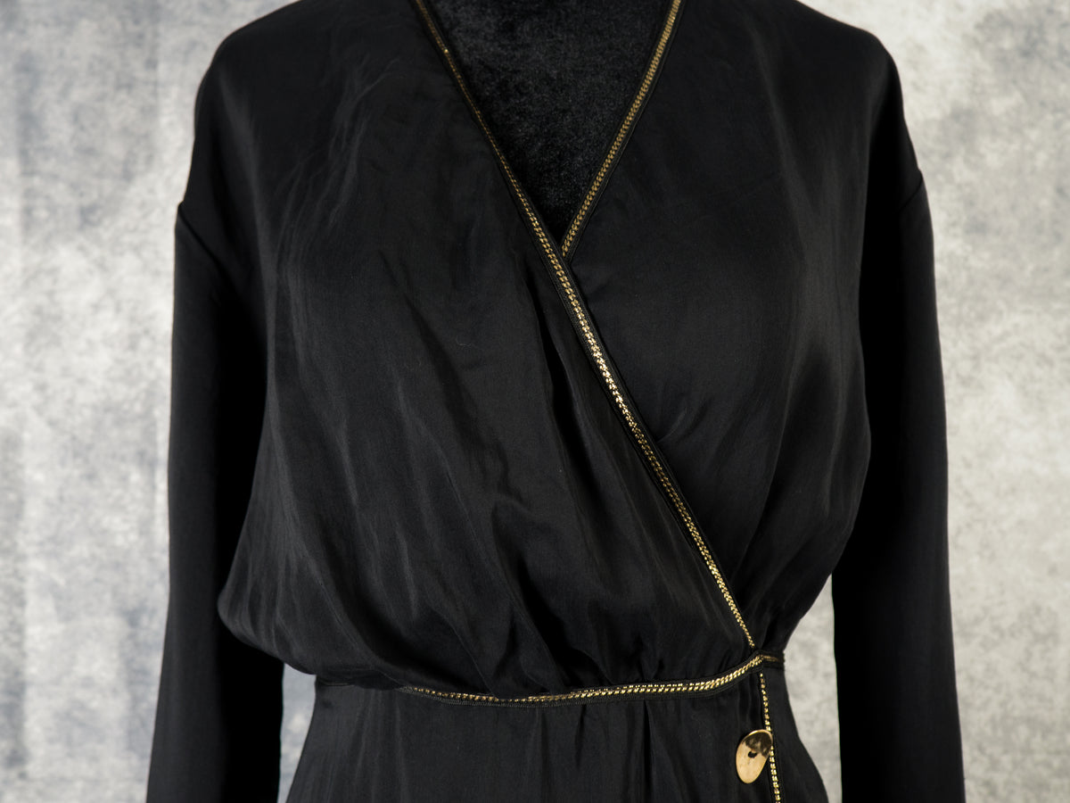 A close-up shot focusing on the bodice of a black faux-wrap dress on a mannequin. It features a deep V-neckline with a draped crossover front and a gathered metallic gold trim. A delicate, metallic gold trim runs along the entire edge of the neckline and the waistline, with a round gold button visible near the hip.