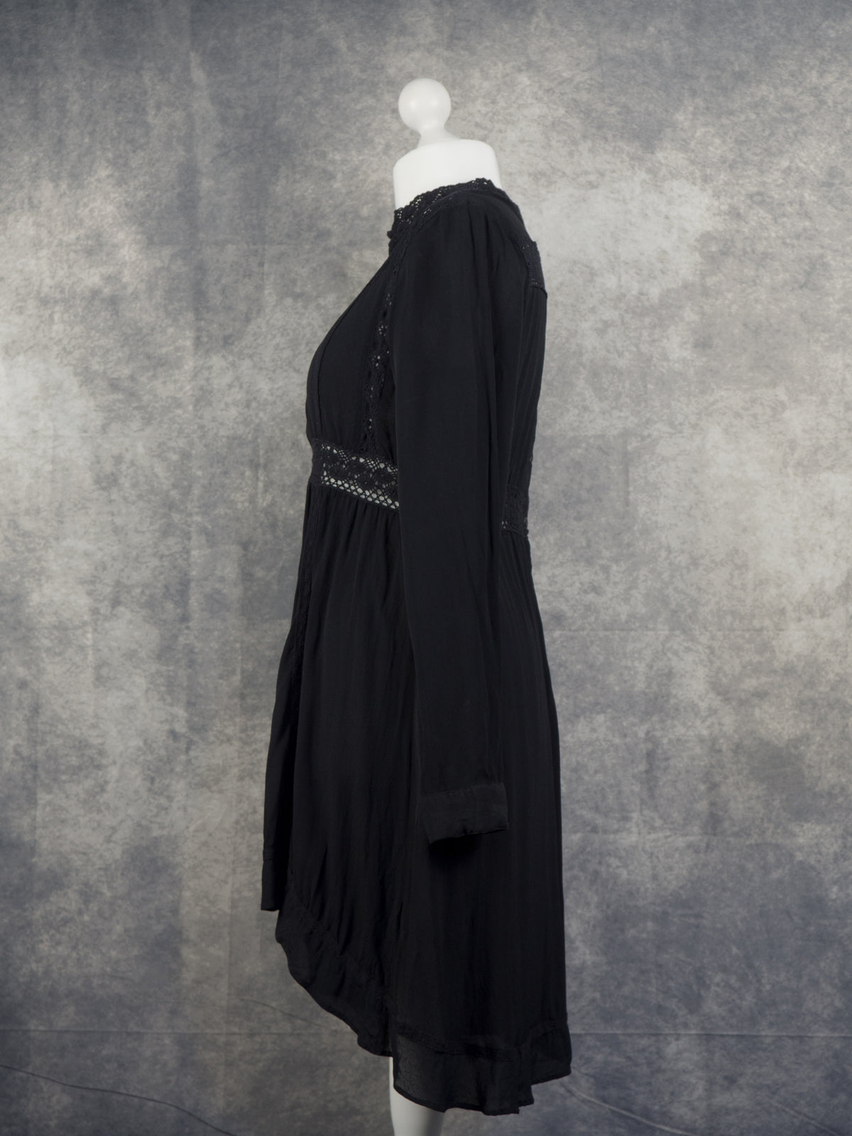 A side profile view of a long-sleeved black mini smock dress displayed on a white mannequin. The dress features an empire waist with a semi-sheer crocheted lace insert panel and subtle lace detailing along the high neckline. The lightweight fabric drapes into a slightly gathered hemline, set against a textured grey studio background.