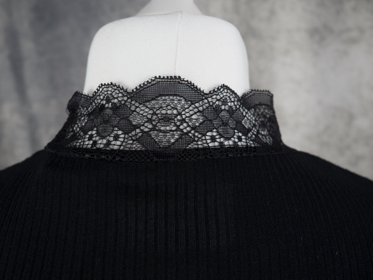 A close-up of a black ribbed long-sleeve knit top on a white mannequin torso, showing a wide black lace collar with a scalloped edge against a gray textured background.