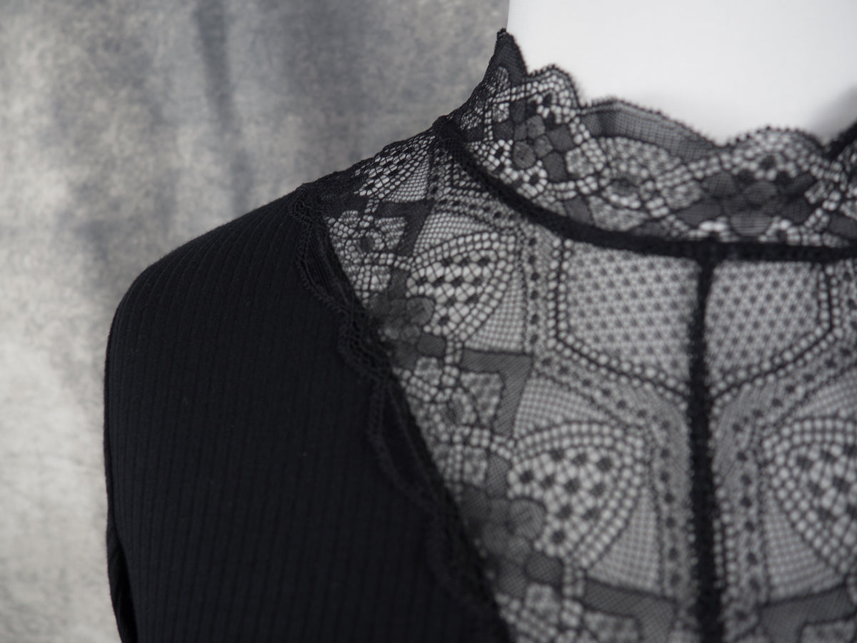 Detail of a black ribbed long-sleeve top with a sheer black lace V-neck panel, displayed on a white mannequin torso against a textured gray background.