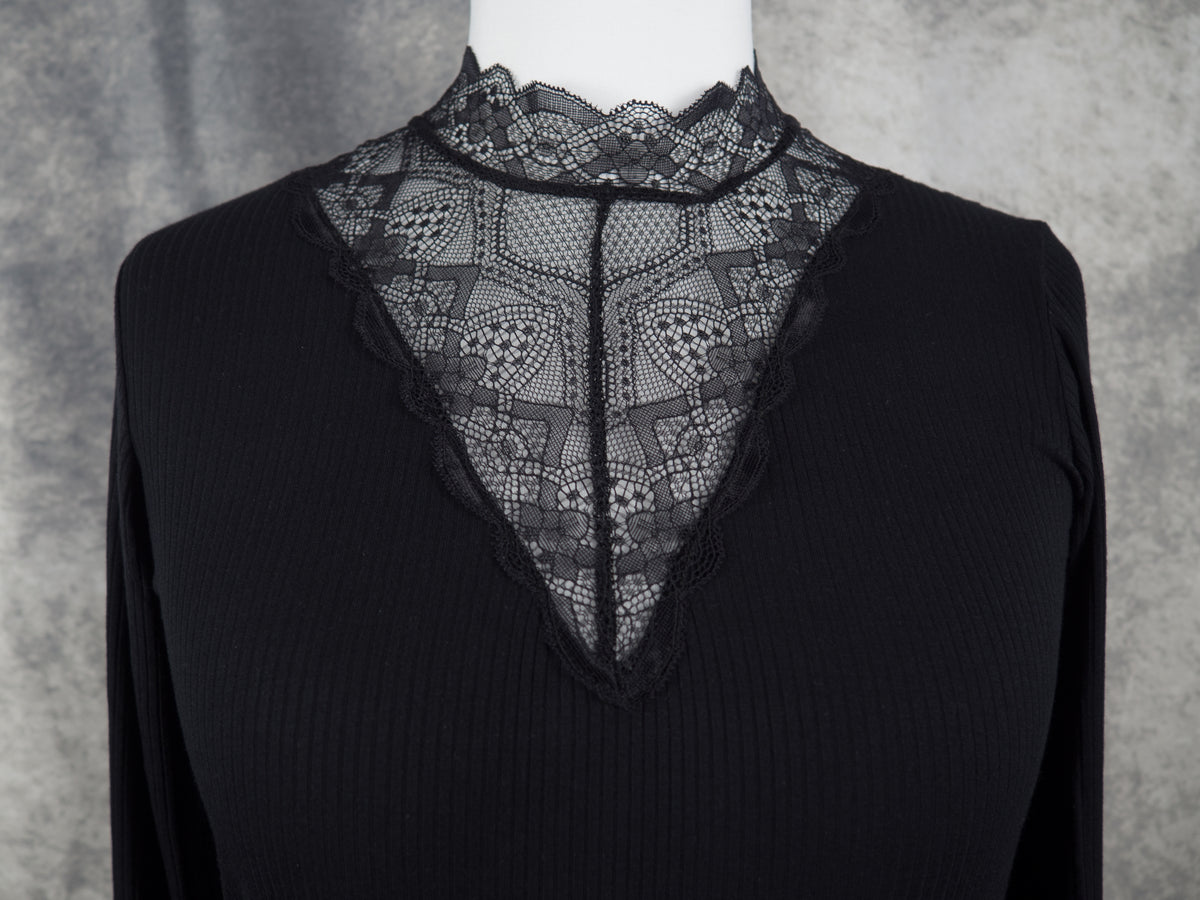 A black ribbed long-sleeve top with a sheer black lace V-neck panel, displayed on a white mannequin torso against a textured gray background.