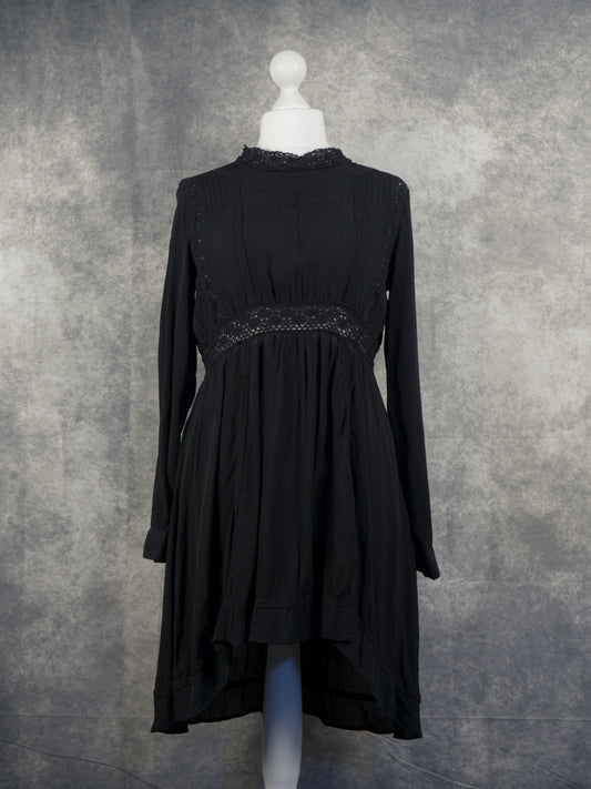 A full front view of a long-sleeved black mini smock dress displayed on a white mannequin. It features a high, lace-trimmed collar and a prominent, semi-sheer crocheted lace panel running horizontally across the empire waist. The lightweight fabric drapes into a tiered, high-low hemline that is shorter in the front, set against a textured grey studio background.