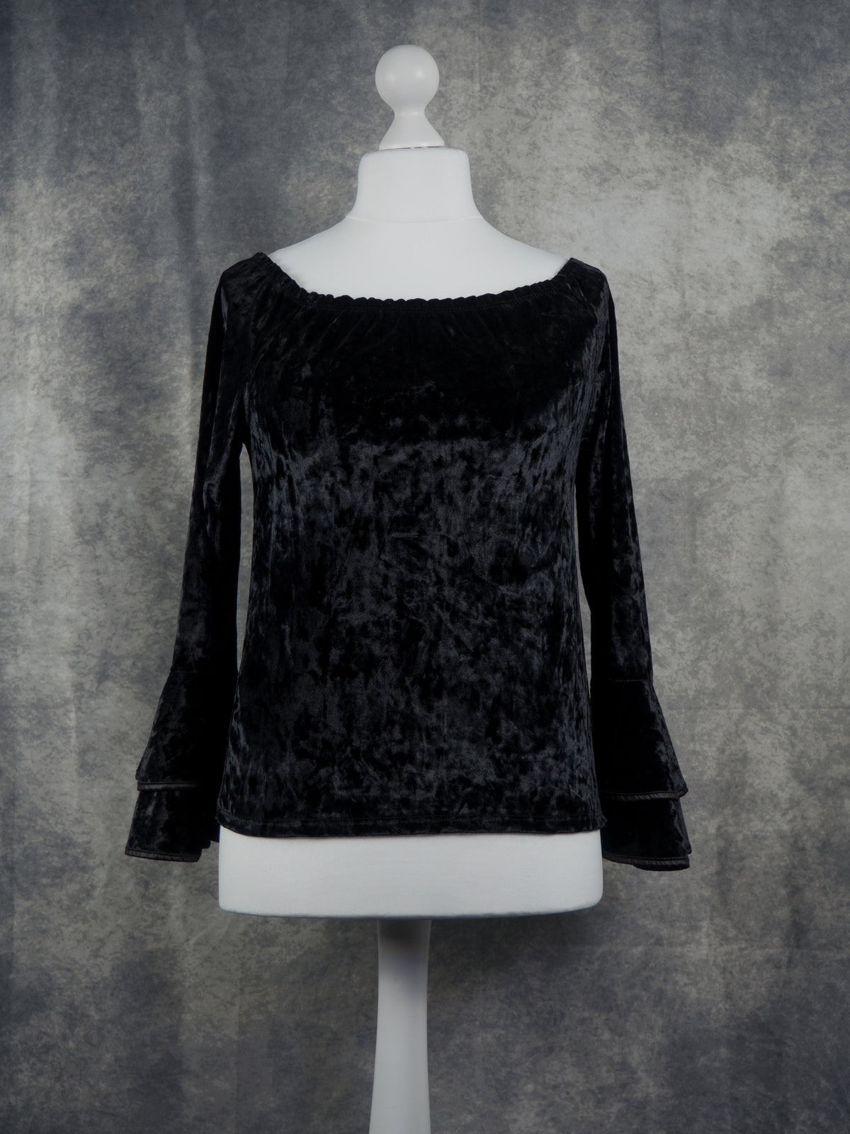 A black velvet long-sleeve top with a wide boat neckline and flared bell sleeves, displayed on a white mannequin torso against a textured gray backdrop.