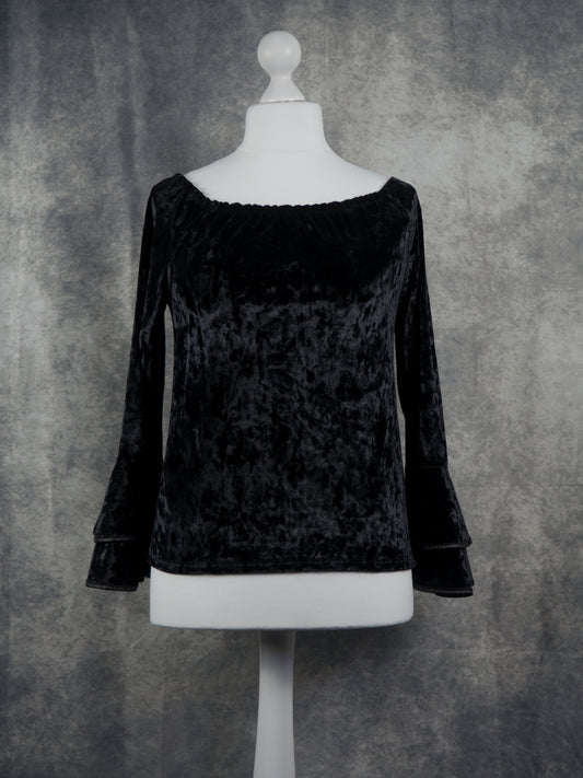 A black velvet long-sleeve top with a wide boat neckline and flared bell sleeves, displayed on a white mannequin torso against a textured gray backdrop.