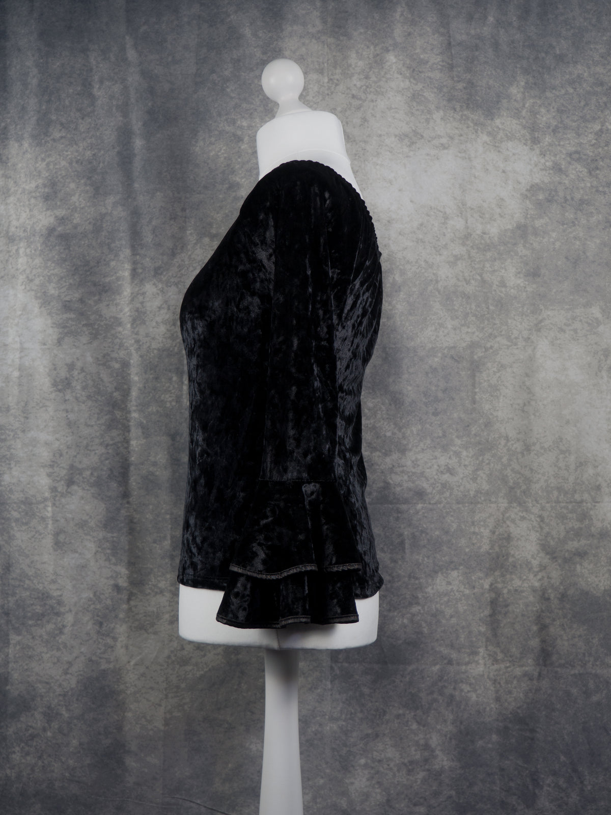 A black velvet longline top or dress displayed on a white mannequin torso against a textured gray studio backdrop.