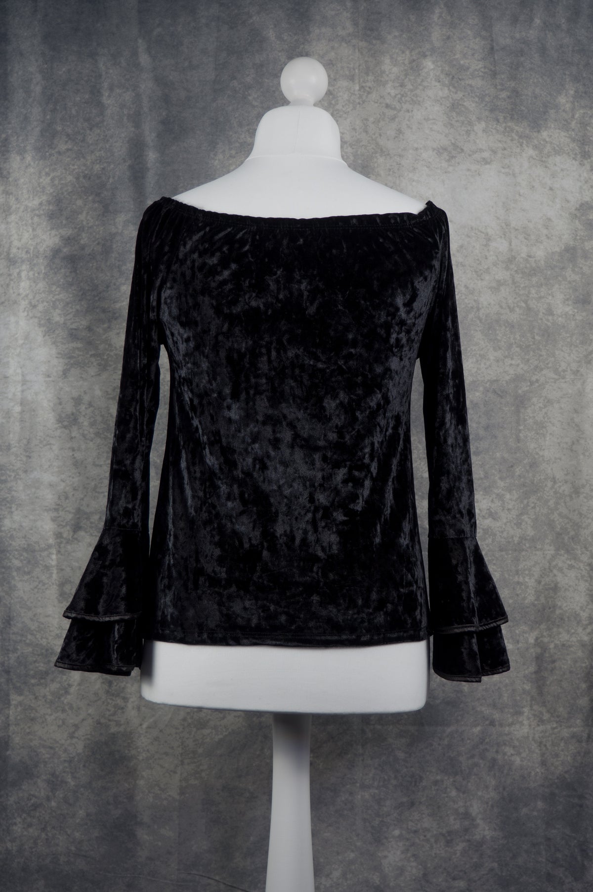 A black velvet off-the-shoulder long-sleeve top with flared bell sleeves, displayed on a white mannequin torso against a textured gray studio backdrop.