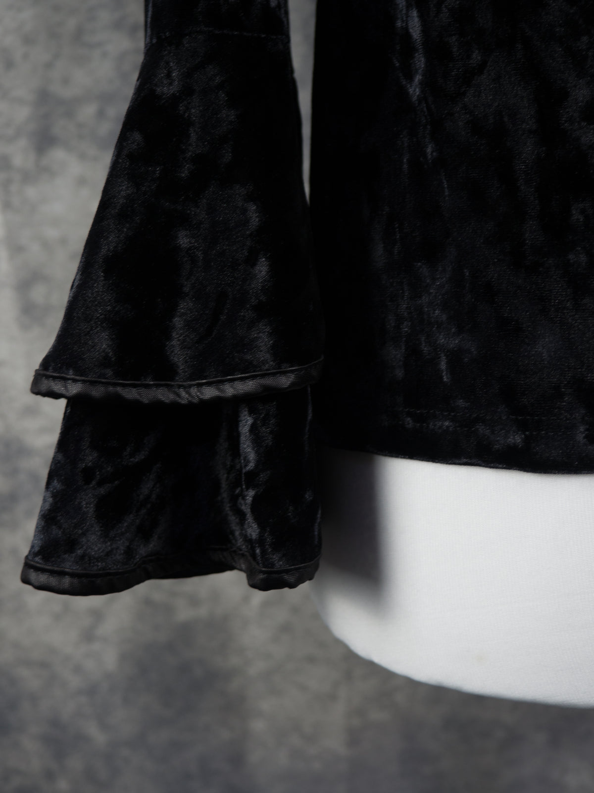 Close-up of a black velvet garment with layered ruffle cuffs, shown on a white mannequin against a textured gray background.
