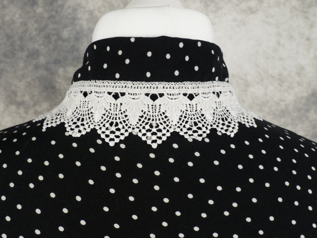 Close-up of the back of the collar on a black garment with a white polka dot pattern. The collar is detailed with a wide, ornate band of white crochet lace trim featuring a delicate scalloped edge that rests across the shoulders. The background is a textured grey wall.