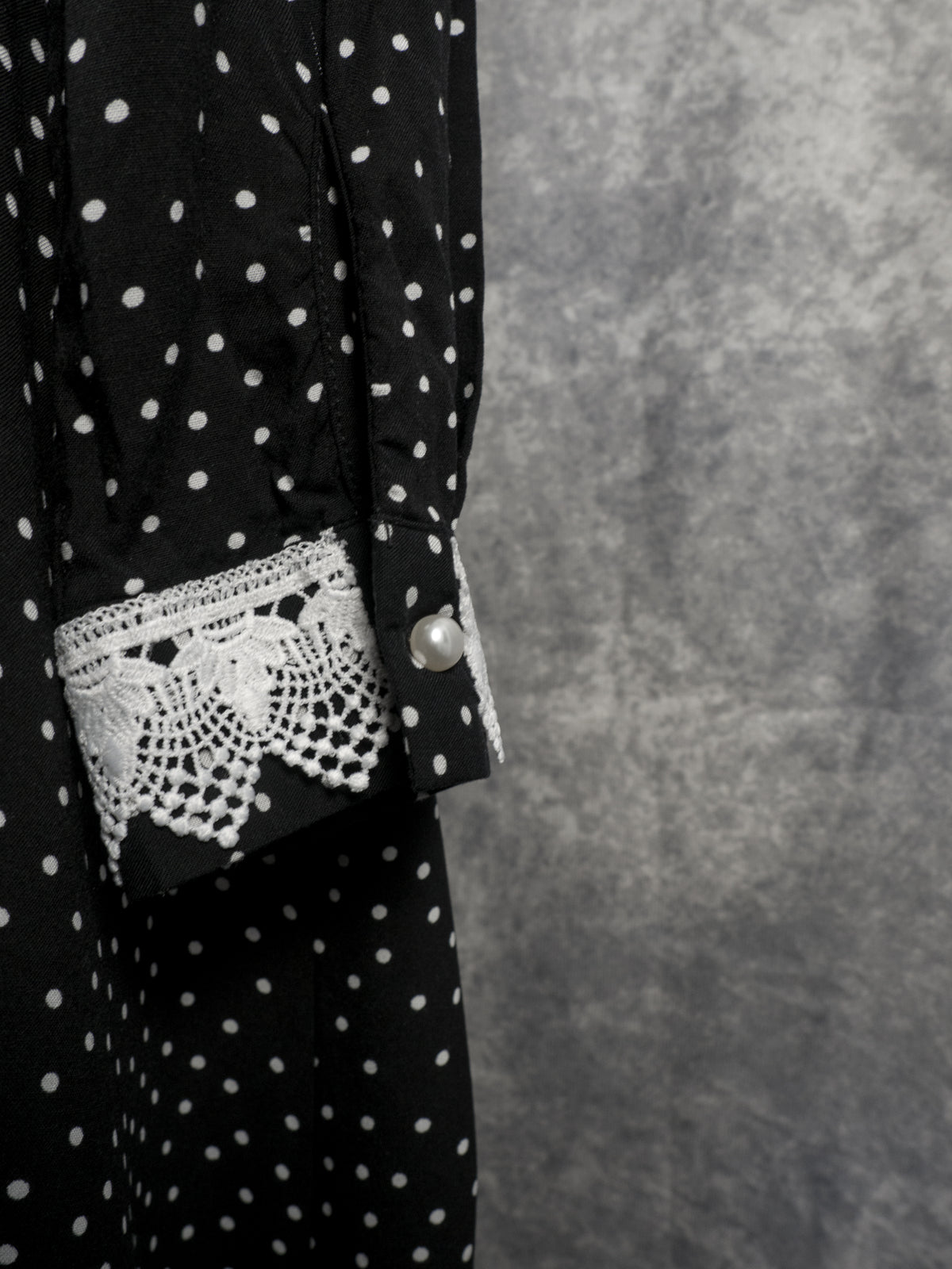 Close-up of the sleeve cuff of a black garment with a white polka dot pattern. The cuff is fastened with a round white pearl-like button and detailed with a patch of intricate white crochet lace. The background is a textured grey wall.
