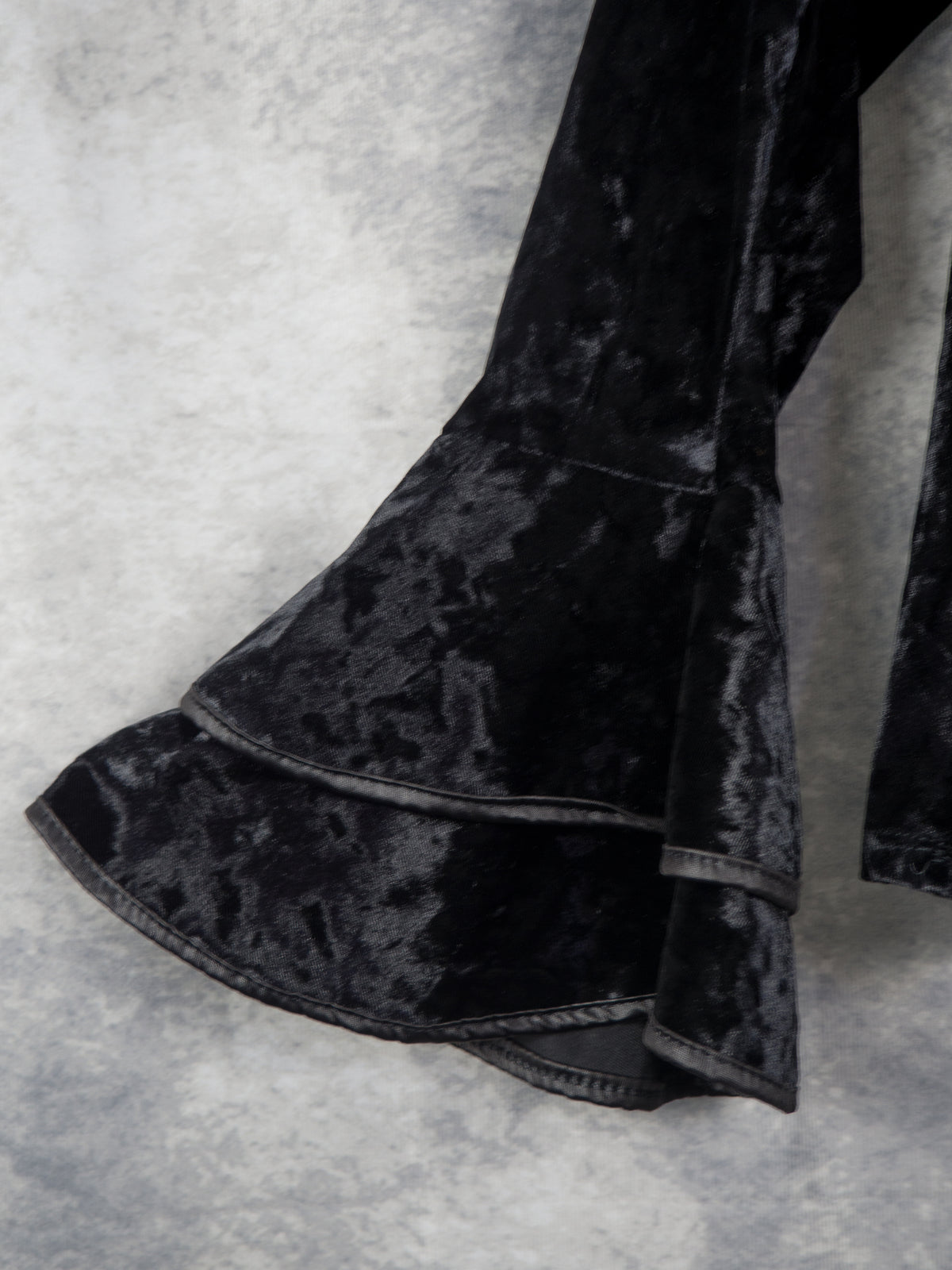Close-up of a black velvet garment with a flared ruffled hem, shown against a textured gray background.