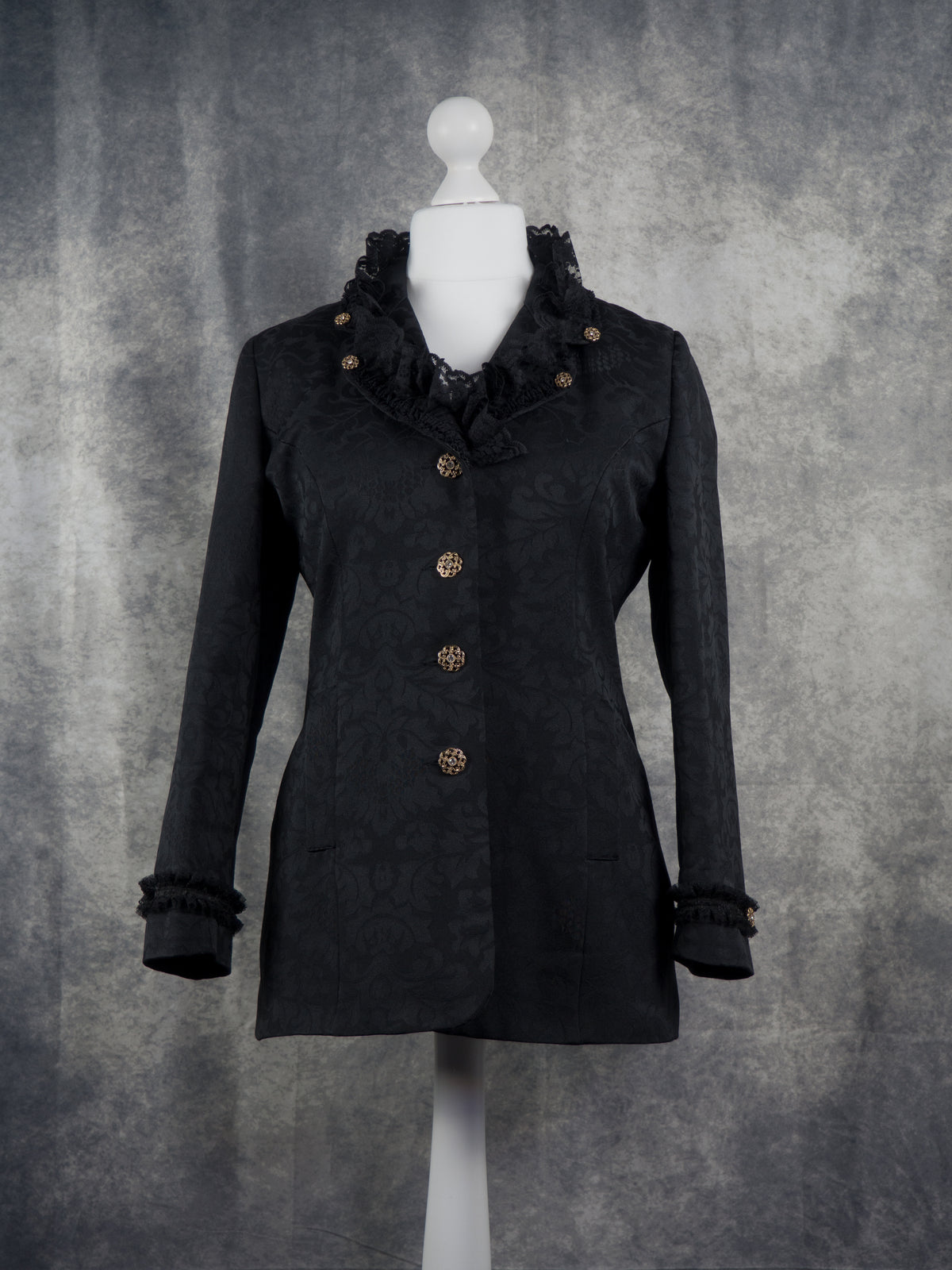 Front view of a black long-sleeve blazer with a textured jacquard pattern, ruffled collar, and decorative gold buttons, displayed on a white mannequin torso against a mottled gray studio backdrop.
