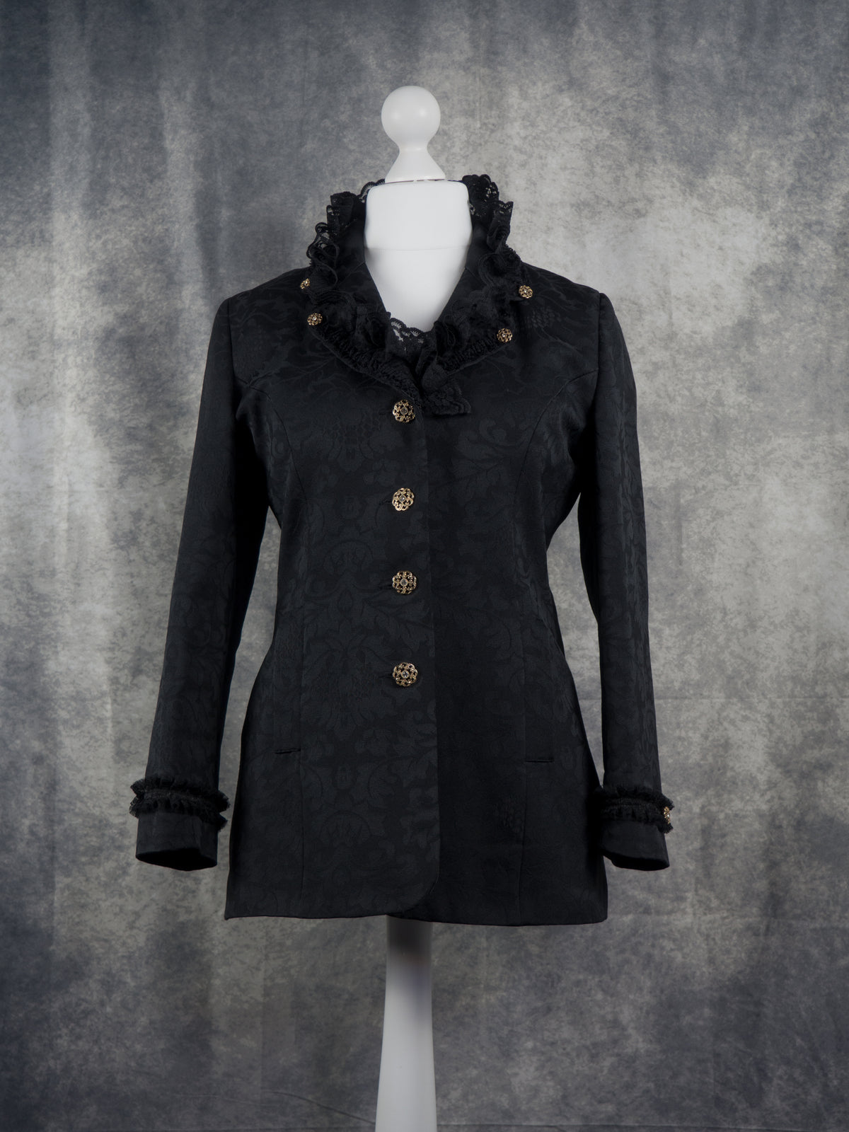 Front view of a black long-sleeve jacket with ornate ruffled trim at the collar and cuffs, featuring decorative gold-toned buttons, displayed on a white mannequin torso against a textured gray backdrop.