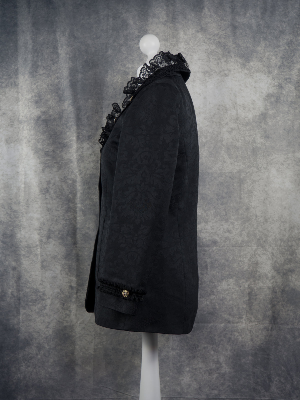 Side view of a black textured blazer with a prominent black lace ruffle collar, displayed on a white mannequin torso against a mottled gray studio backdrop.