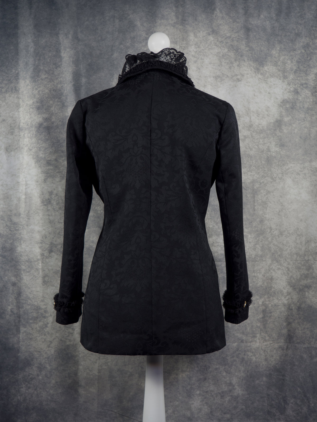 Back view of a black long-sleeve coat with a high lace-trimmed collar and ruffled cuffs, displayed on a mannequin against a textured gray backdrop.