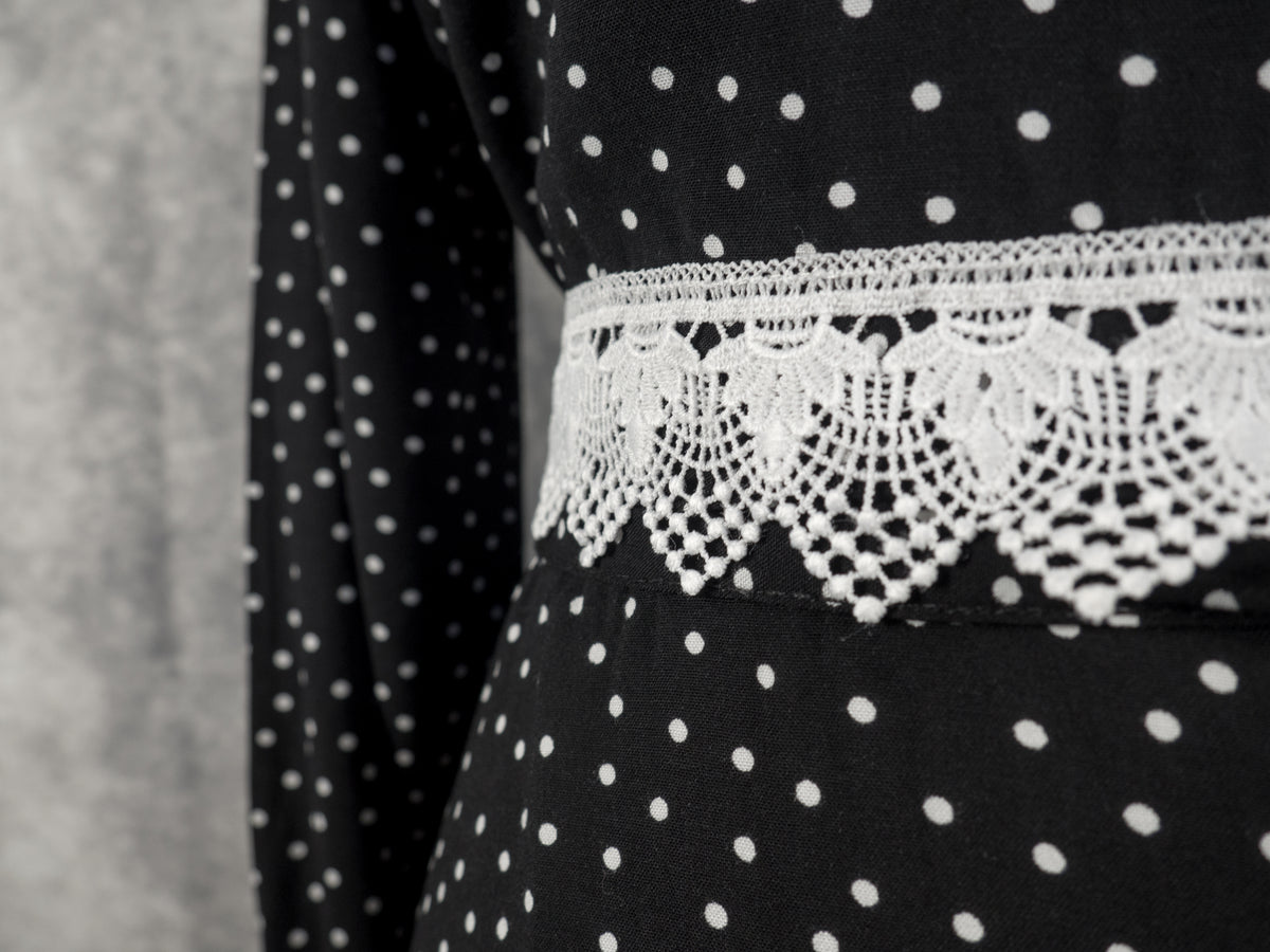 Close-up of a black dress with white polka dots and a wide white lace trim around the waist, shown against a neutral gray background.