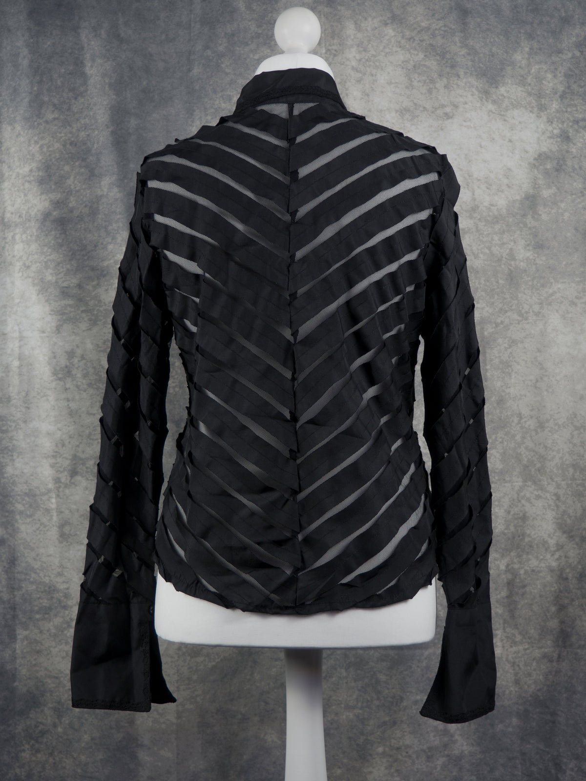 A black long-sleeve blouse with a high collar and a unique chevron, or zigzag, cutout pattern on the bodice and sleeves, displayed on a white mannequin torso against a textured gray background.