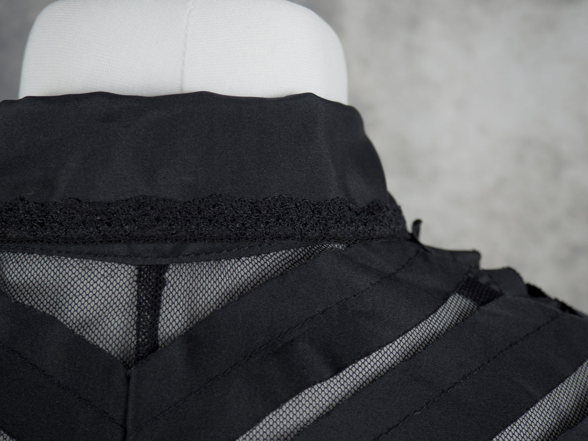 A close-up, detailed shot focusing on the back collar and upper back of the black blouse on the mannequin. It highlights a narrow strip of black lace trim running horizontally along the base of the solid collar. Below the lace, the alternating diagonal strips of solid black fabric and sheer mesh meet at a central vertical seam, creating a downward V-shaped chevron pattern.