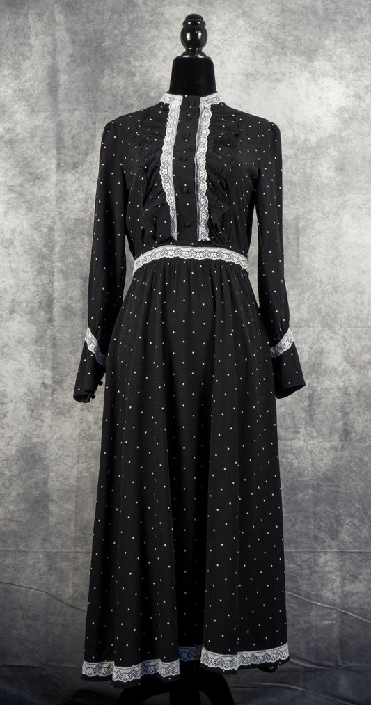 A full-length front view of the black vintage-style maxi dress, displayed on a black mannequin against a textured grey background. The dress features a repeating pattern of small white stars across the entire fabric. It is styled with distinct vertical ruffles on the chest, flanked by white lace trim that runs along the high collar down to the buttoned front placket. Matching white lace accents the waistband, the cuffs of the long sleeves, and the bottom hemline, creating a complete, prairie-inspired look.