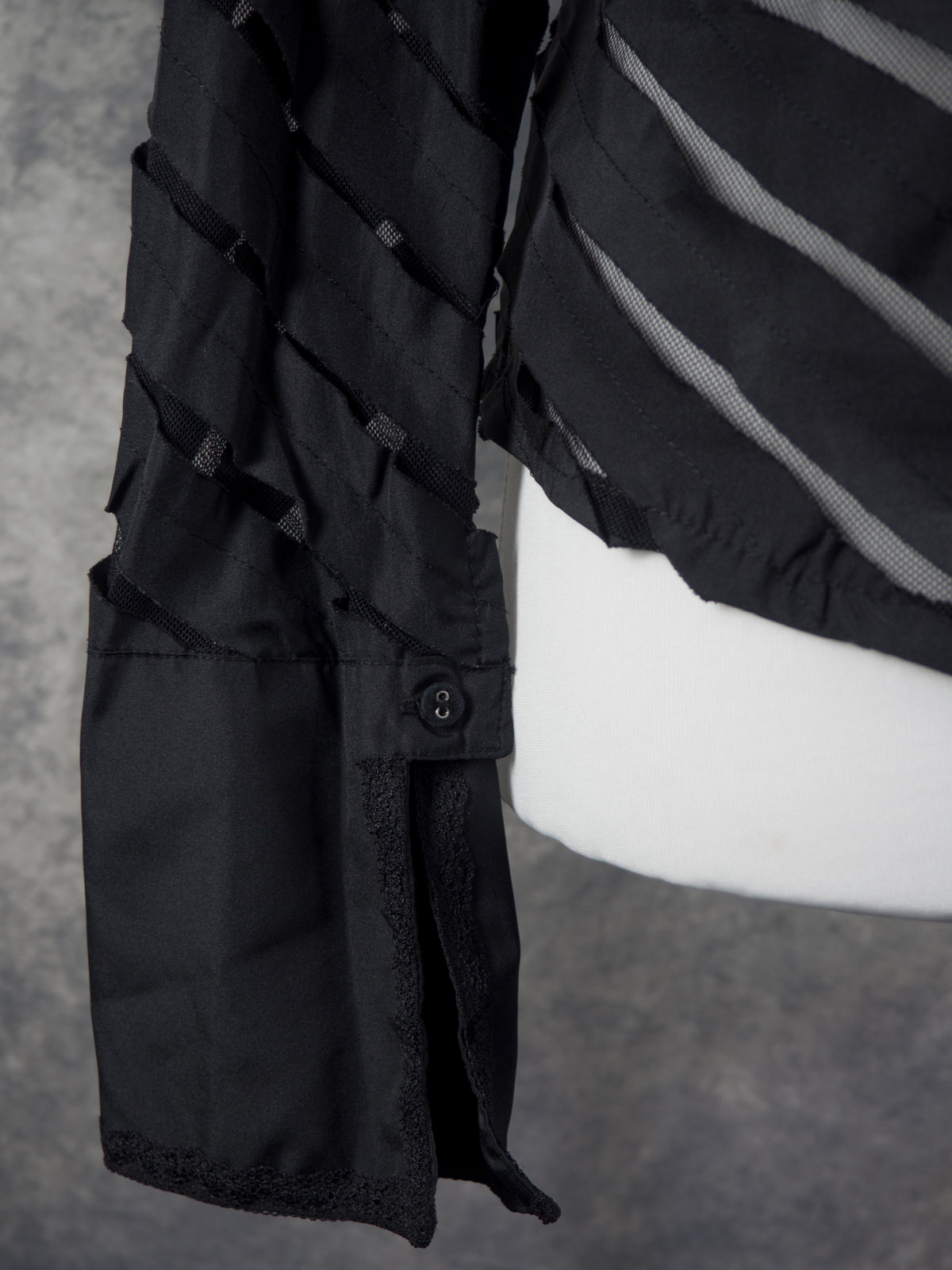 A close-up view showing the lower sleeve, cuff, and side hem of the black blouse on the mannequin. The sleeve features the same alternating diagonal panels of solid black fabric and sheer mesh. It ends in a wide, solid black cuff with a small button fastening at the slit, which is lined with a delicate black lace trim along the edges.