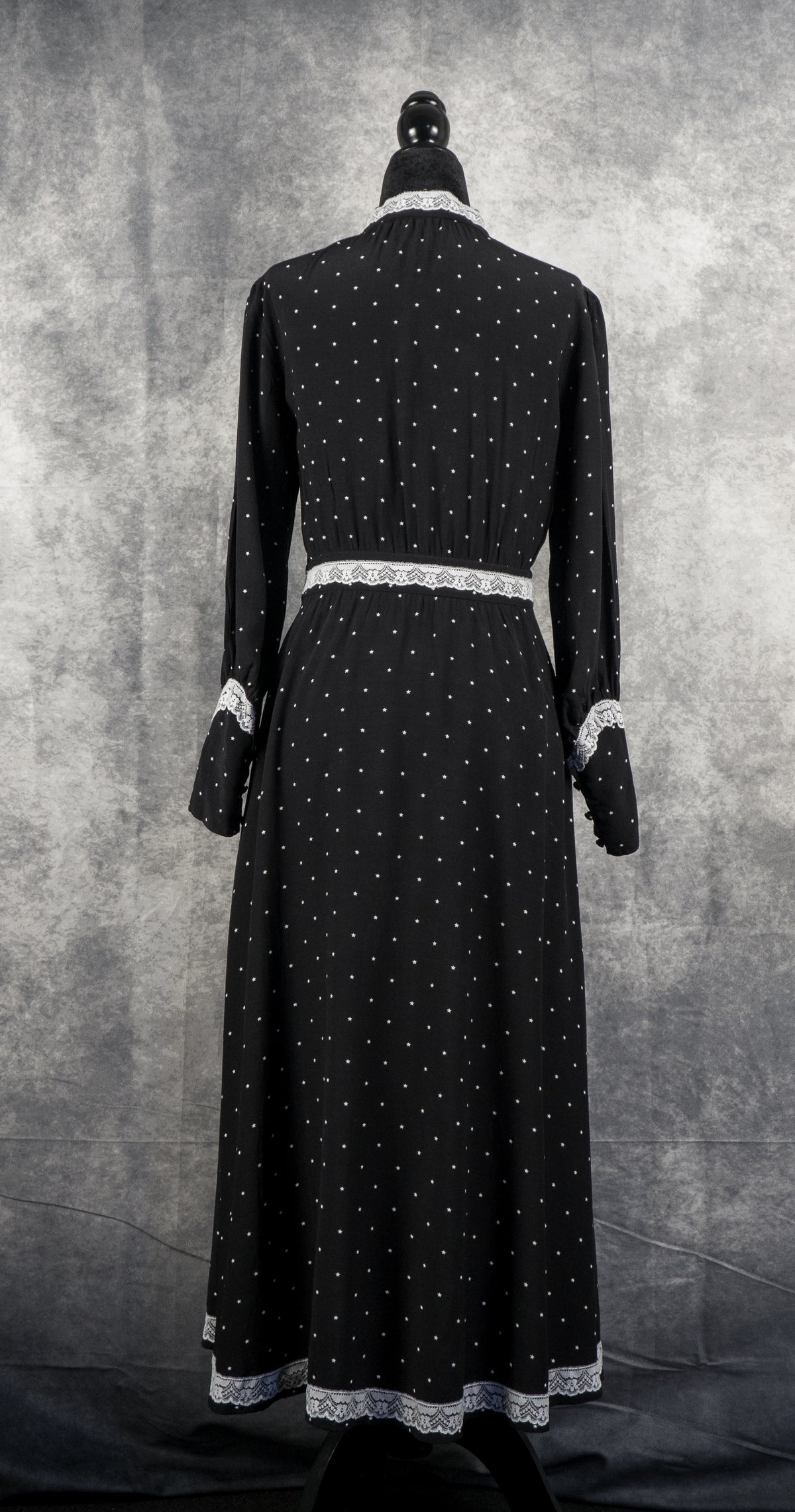 A full-length back view of the black vintage-style maxi dress, displayed on a black mannequin against a textured grey background. The dress showcases its uniform pattern of small white stars covering the back bodice, long sleeves, and a flowing a-line skirt. It features white lace trim accenting the high collar, the defined waistband, the cuffs of the long sleeves, and the entire bottom hemline.