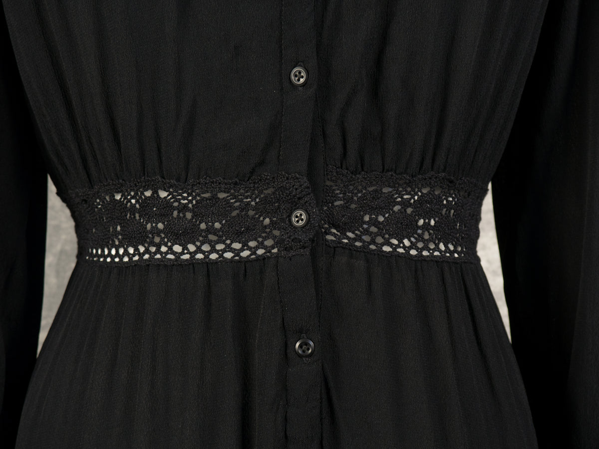 A close-up view of the back waistline of a black dress, highlighting a semi-sheer crocheted lace insert panel that features an intricate geometric design. Small black buttons run vertically along the center placket, intersecting the lace band and adding a delicate detailed finish against the soft fabric.