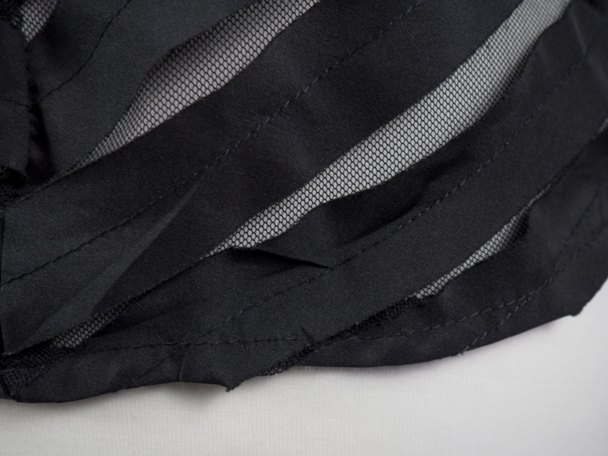Close-up of a black sheer mesh fabric with wide, layered ruffle-like strips and visible stitching, laid against a light neutral background.