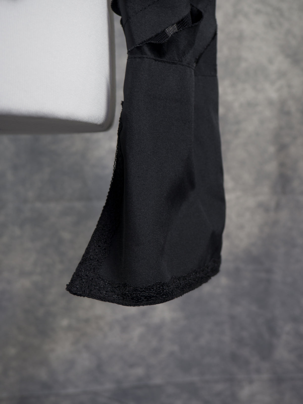 A close-up view showing the lower sleeve, cuff, and side hem of the black blouse on the mannequin. The sleeve features the same alternating diagonal panels of solid black fabric and sheer mesh. It ends in a wide, solid black cuff with a small button fastening at the slit, which is lined with a delicate black lace trim along the edges.