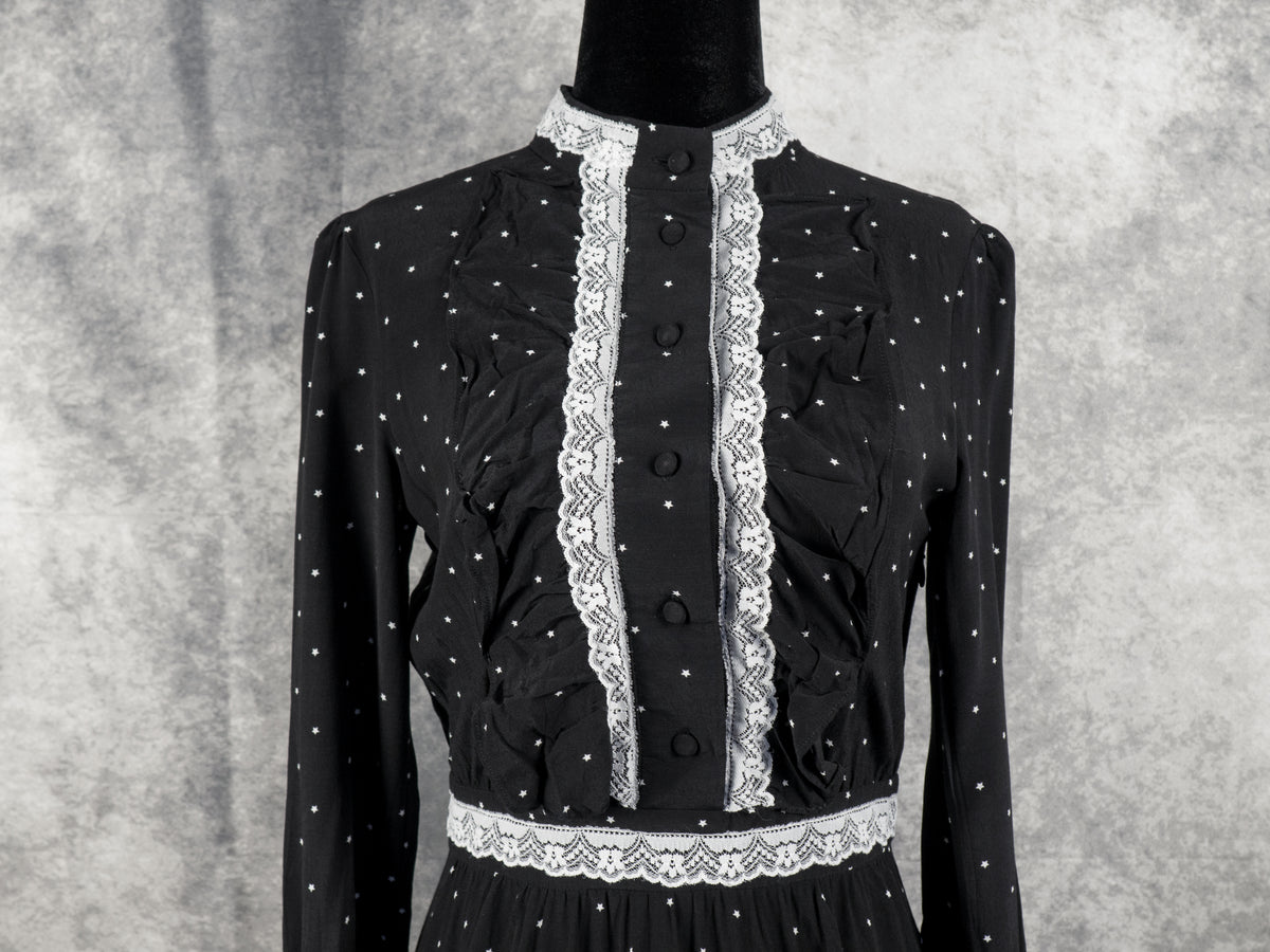 A medium close-up front view focusing on the upper bodice of the black vintage-style dress. The image highlights the high collar and vertical placket lined with detailed white lace trim and five black fabric-covered buttons. Textured vertical ruffles flank the central lace detail on the chest, all set against the black fabric patterned with small white stars. The lace-trimmed waistband is visible at the bottom against the grey backdrop.