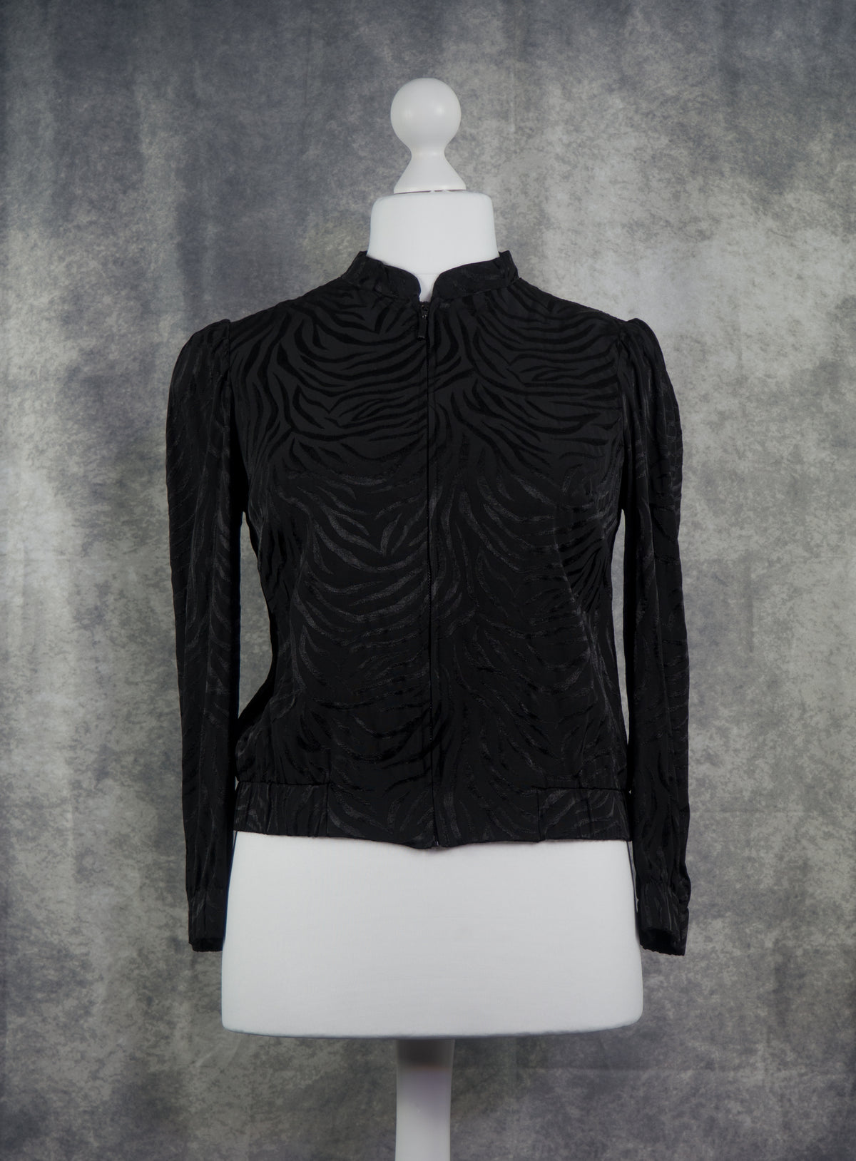 A black long-sleeve cropped jacket with a textured, wavy embossed pattern, displayed on a white mannequin torso against a mottled gray background.