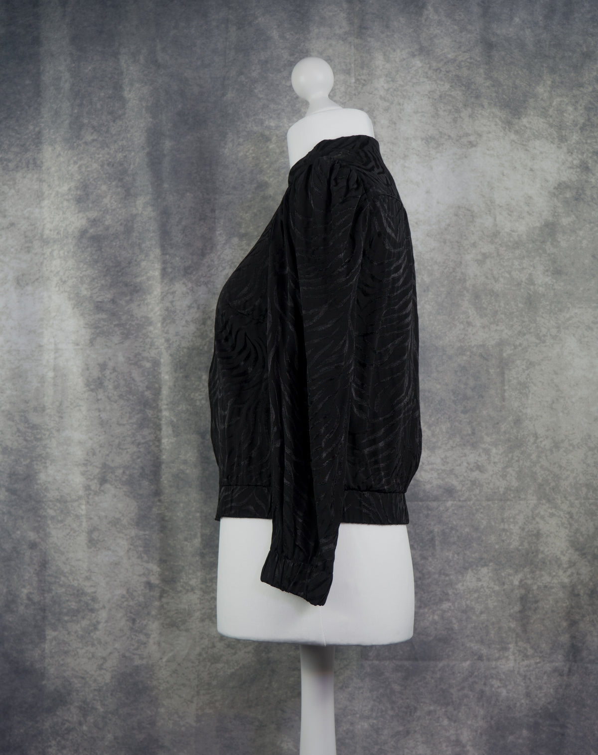 A black textured long-sleeve blouse with a gathered, balloon-style silhouette, displayed on a white mannequin torso against a mottled gray studio backdrop.