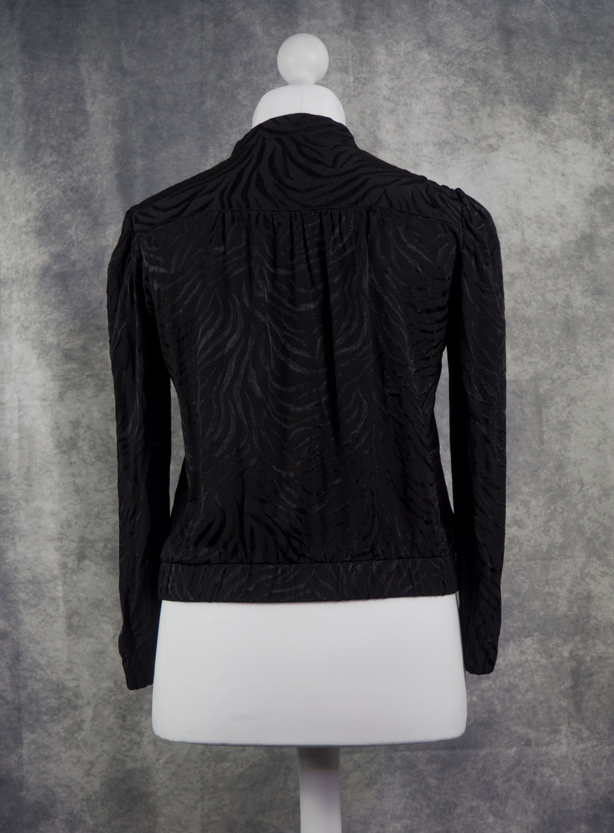 Black long-sleeve cropped jacket with a textured, wavy jacquard pattern, displayed on a white mannequin torso against a textured gray studio backdrop.