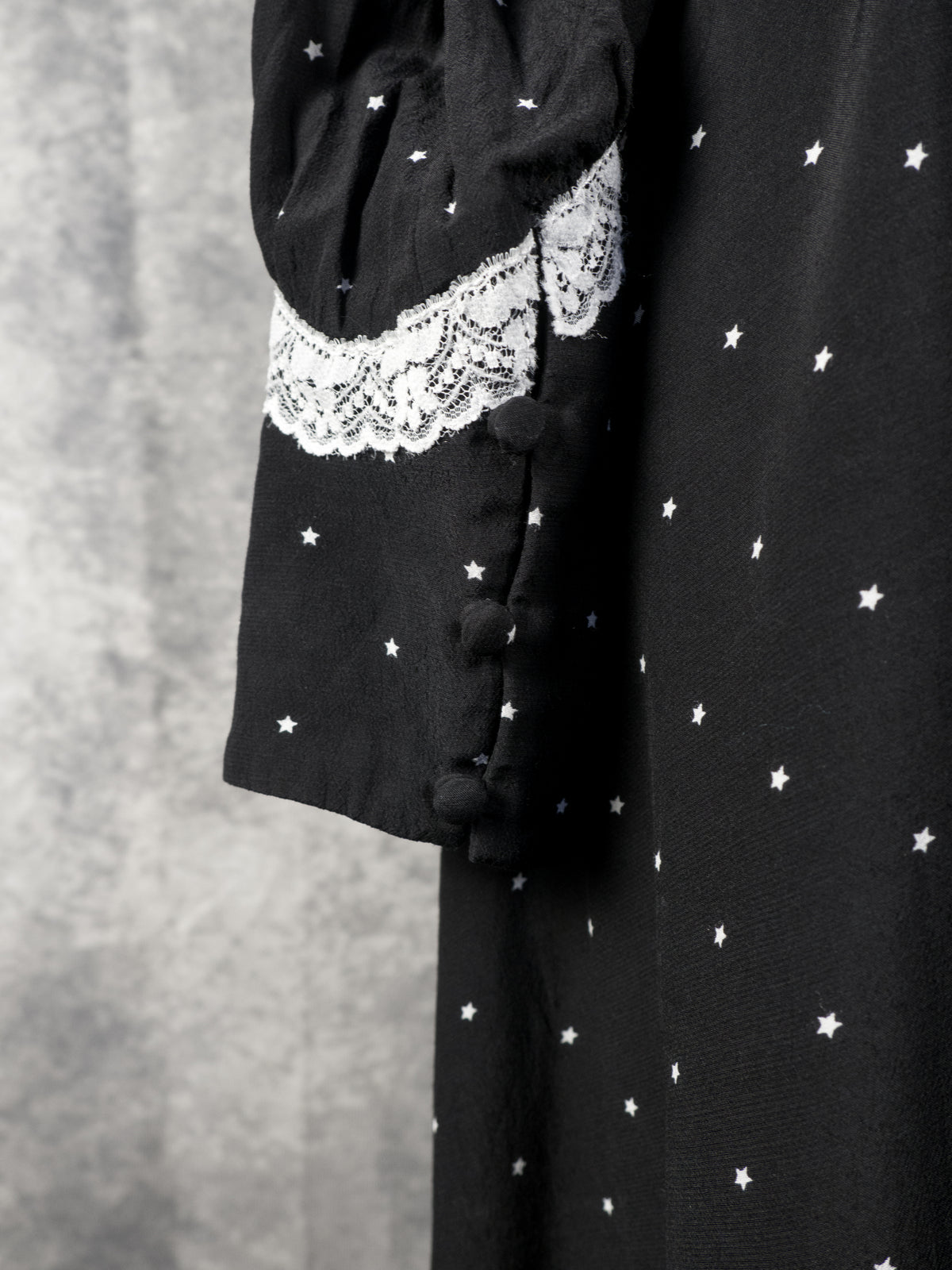 A close-up shot focusing on the sleeve cuff of the black vintage-style dress. The image highlights a band of detailed white lace trim separating the upper sleeve from a pointed cuff extension, which features small black buttons fastened along its edge. The fabric is patterned with the continuous small white stars, set against the textured grey background.