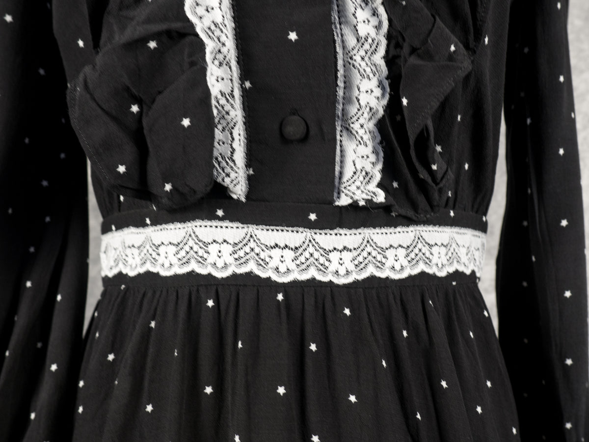 A close-up view focusing on the waist and lower bodice of the black vintage-style dress. The image highlights the horizontal white lace trim along the defined waistband, positioned directly below the buttoned placket and textured ruffles on the chest. The continuous pattern of small white stars covers the gathered fabric above and below the waist, set against a neutral grey background.