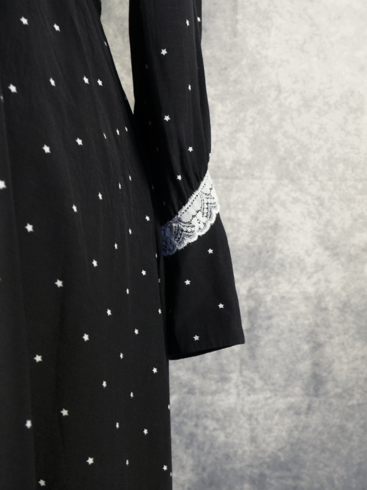 A close-up shot focusing on the sleeve cuff of the black vintage-style dress. The image highlights a band of detailed white lace trim separating the upper sleeve from a pointed cuff extension, which features small black buttons fastened along its edge. The fabric is patterned with the continuous small white stars, set against the textured grey background.