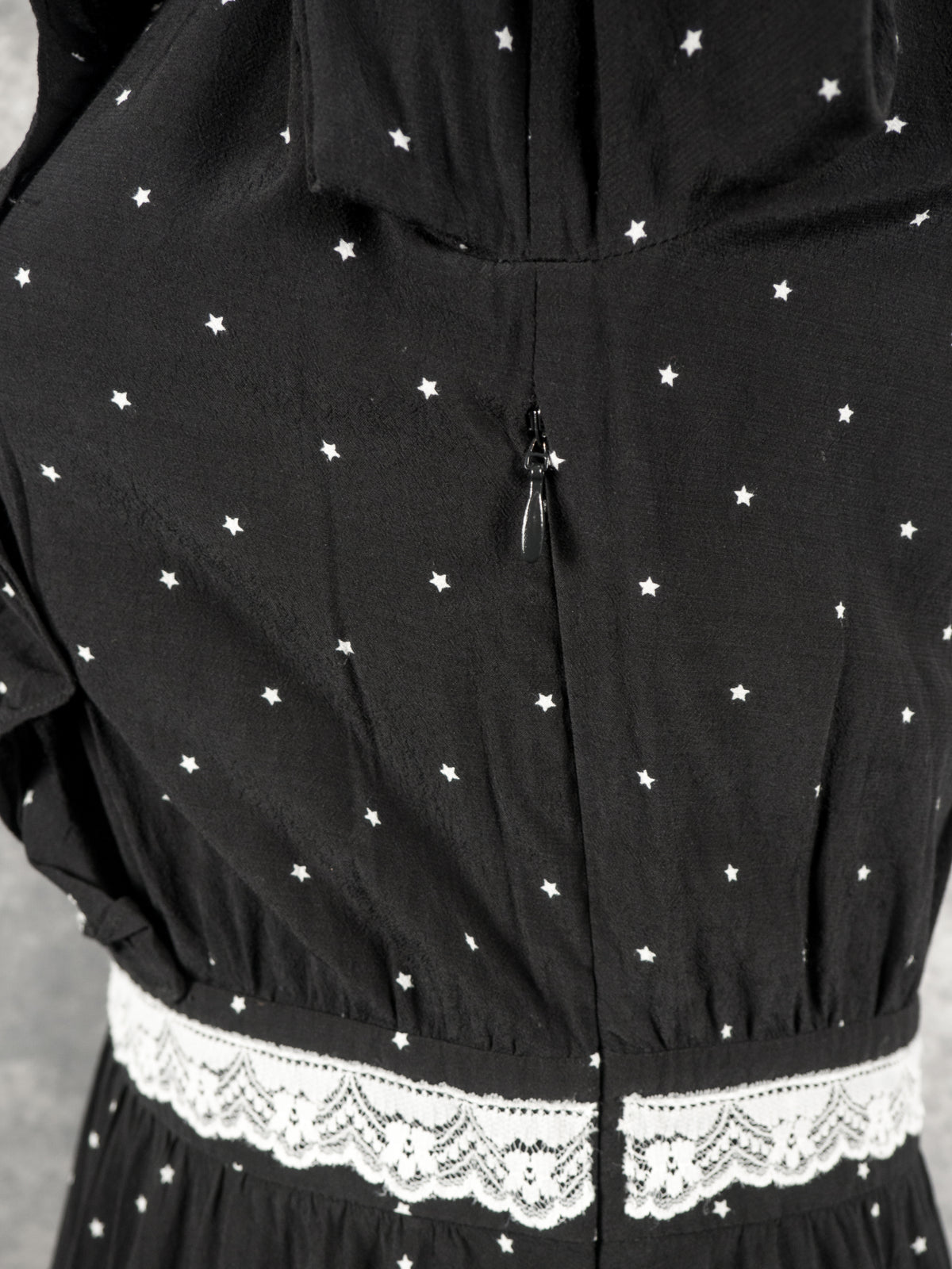 A close-up back view focusing on the upper back zipper closure of the black vintage-style dress. The image reveals a small, black invisible zipper pull located in the center seam of the bodice, just above the gathered white lace waistband. The black fabric is uniformly patterned with the small white stars, set against a soft grey background.