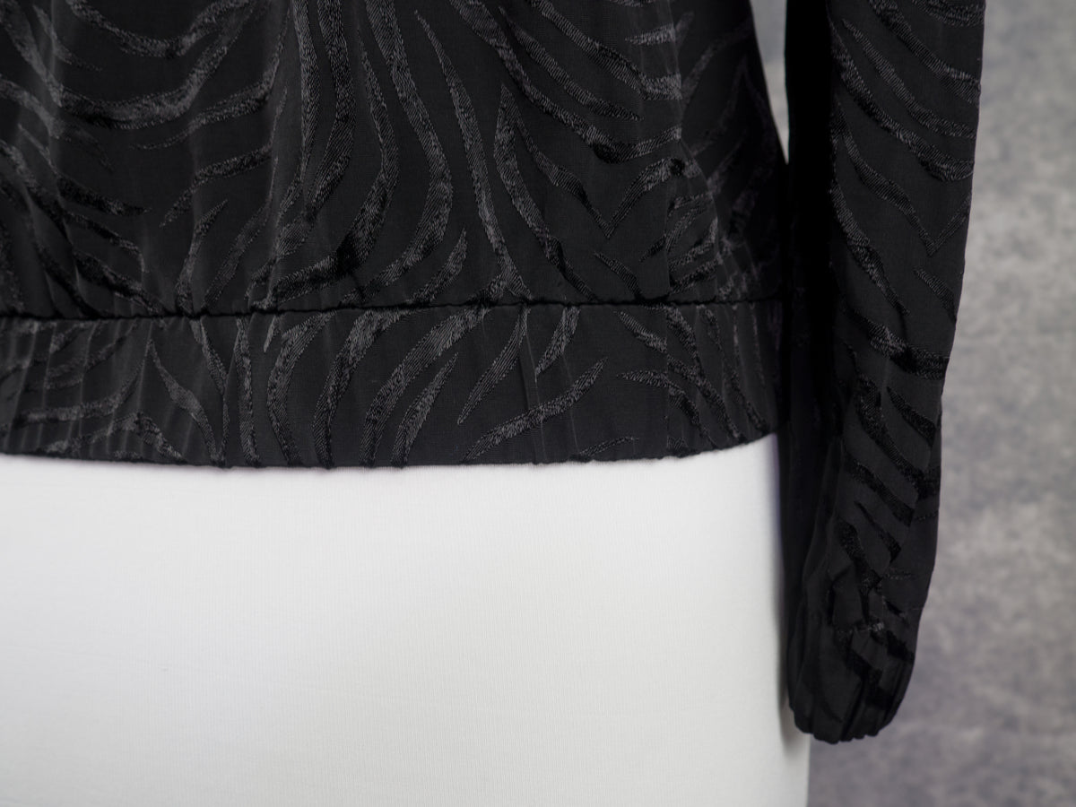 Close-up of a black long-sleeve cropped jacket with a textured, wavy jacquard pattern, displayed on a white mannequin torso.